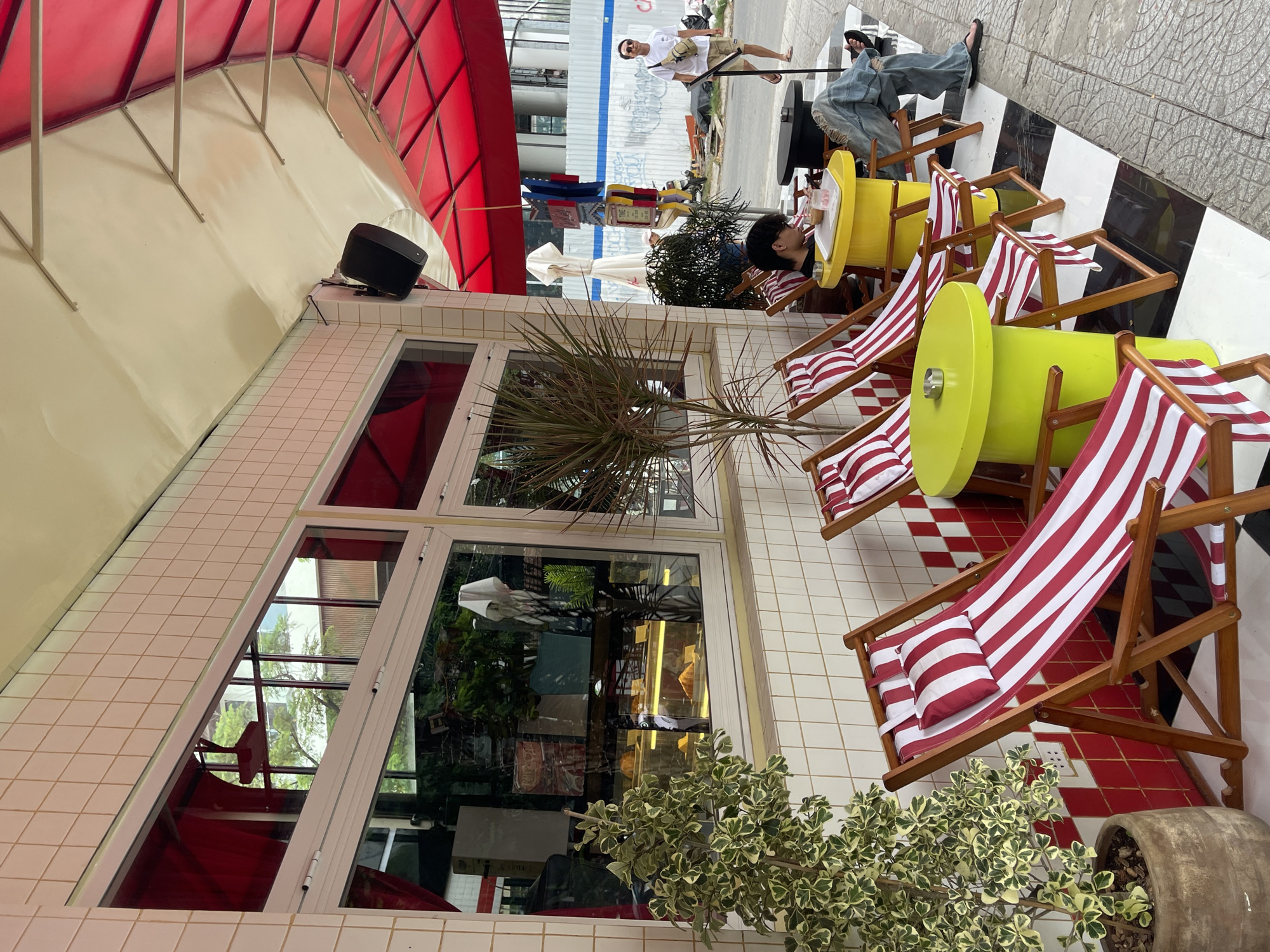 This is an outdoor street-level shot of a cafe in Da Nang, Vietnam. The cafe has a tiled facade in cream and red, with large windows displaying baked goods. Red and white striped deck chairs are set up on a black and white checkered tiled patio, with bright lime green cylindrical tables between them. A person with dark hair wearing a black t-shirt and jeans is seated in one of the chairs, drinking from a cup. Another person in a white t-shirt and shorts is walking past in the background on the sidewalk, which is adjacent to a construction site with a white barrier fence. The cafe is covered by a red awning with a cream-colored underside. A small potted plant with variegated leaves sits to the left of the chairs.