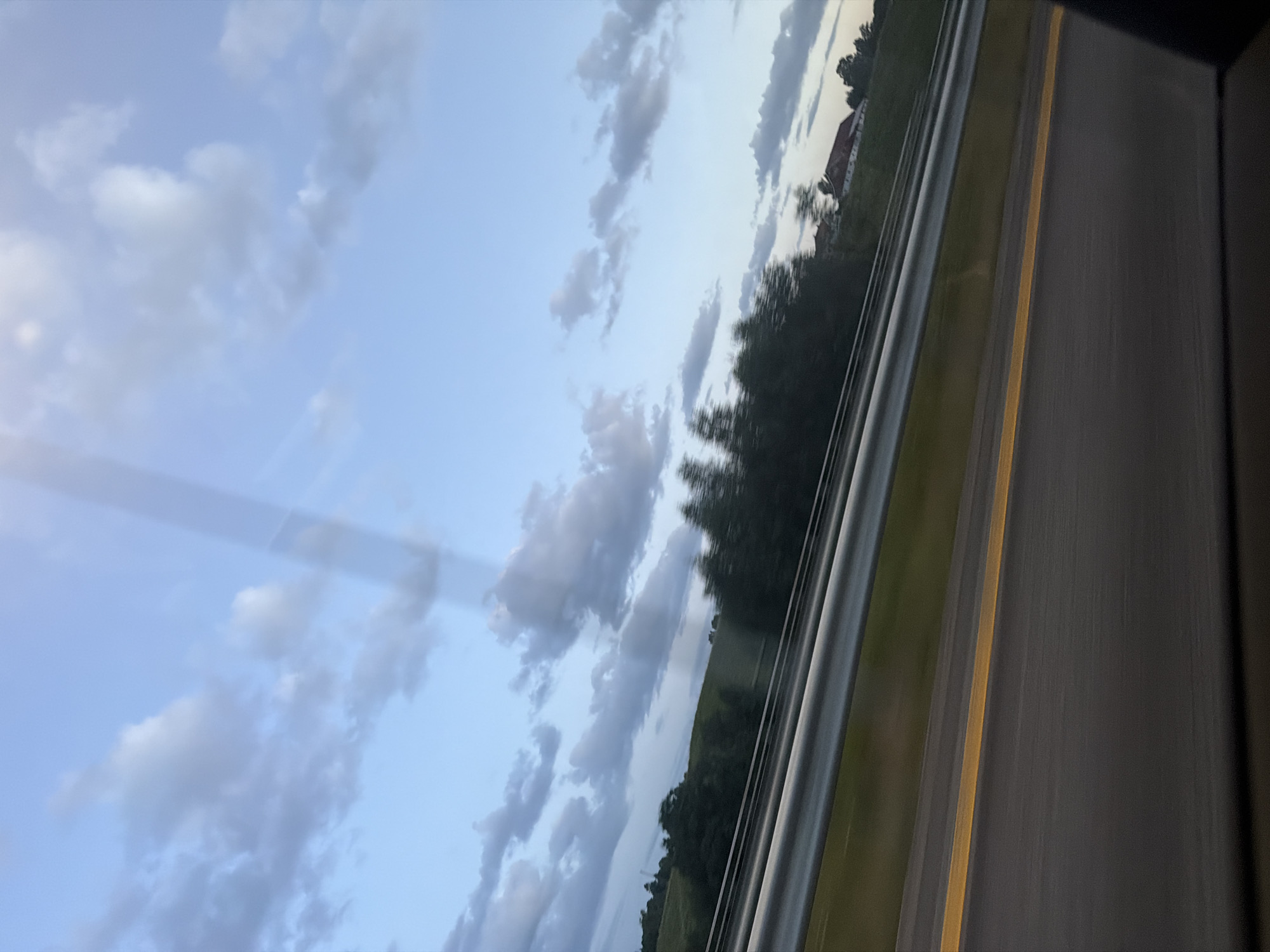 The image shows a view from a moving vehicle, likely a car, on a highway. The foreground features a dark asphalt road with a bright yellow line marking the lane. A silver guardrail runs along the side of the road, separating it from a grassy verge. Beyond the verge, a line of dark green trees is visible, with some appearing blurred due to motion. In the distance, a rolling green hill is topped with a few buildings, one with a red roof. The sky is a pale blue with scattered grey and white clouds, suggesting it may be late afternoon or early evening. There is a faint vertical light streak across the upper portion of the image, possibly from a sunbeam or reflection. The scene appears to be in a rural or suburban area. The provided context specifies Sarpsborg, Norway, suggesting this highway scene is within or near that city.