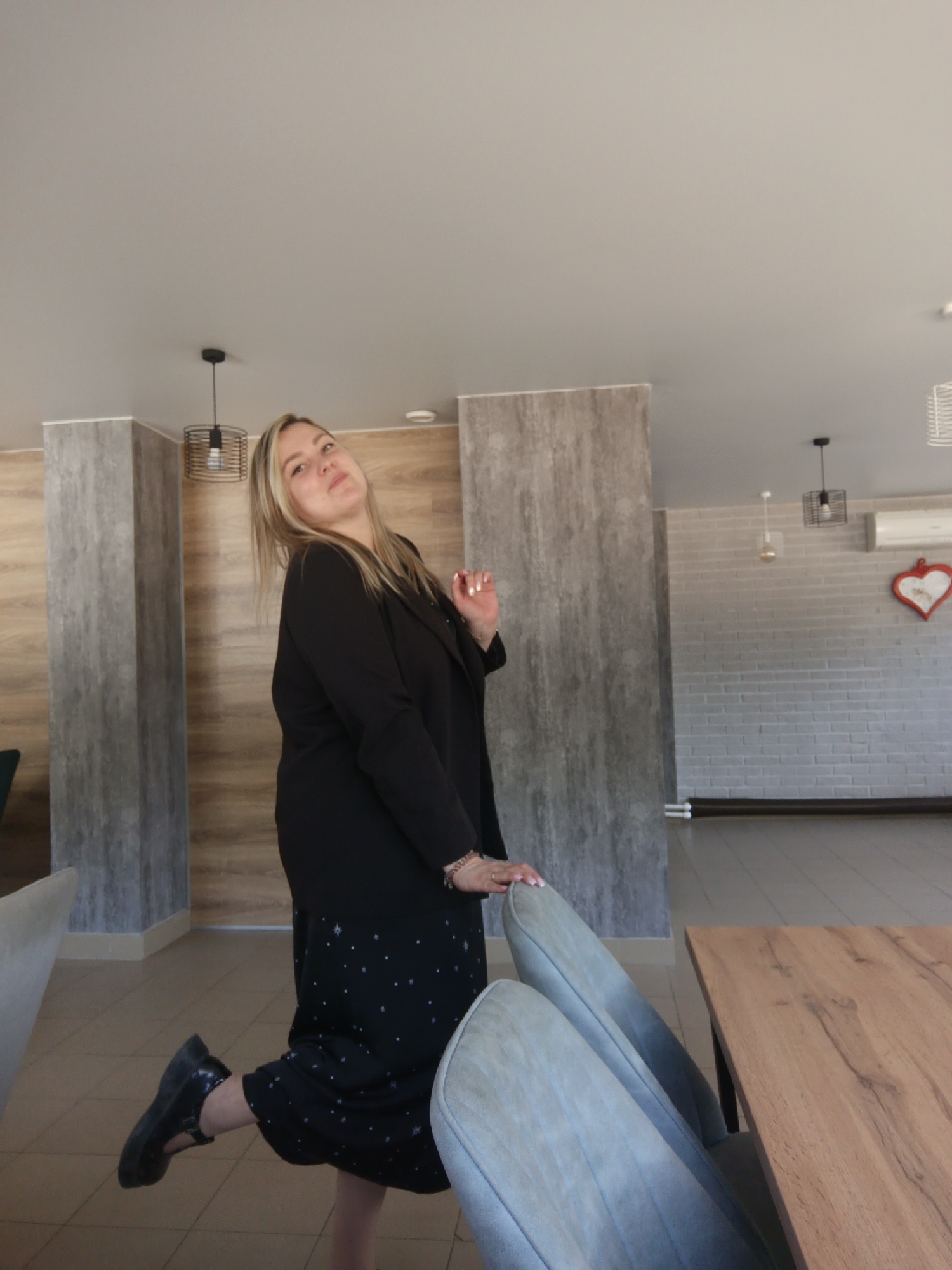 A woman poses in a room with a table and chairs. She wears a black blazer and a dark dress with light-colored stars. Her left leg is bent behind her, and her right foot is planted on the ground. She rests her right hand on the top of a grey chair. The room has decorative panels on the walls, pendant lighting, and a brick wall with a heart decoration. The photograph was taken indoors, with a neutral background. The location is Noviy, Russia.