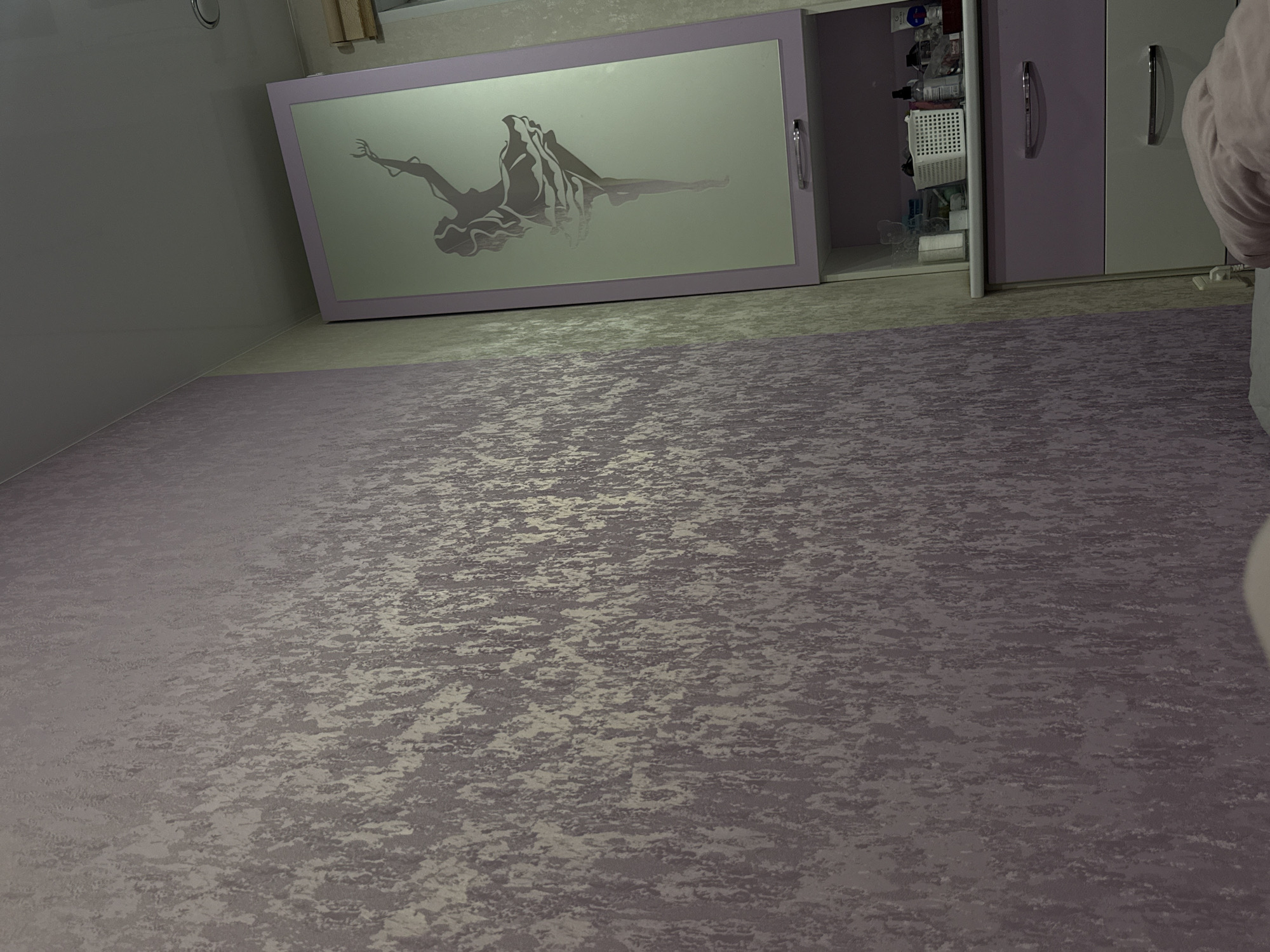 The image displays an interior room, likely a bedroom or dressing area, viewed from a low angle looking across a patterned floor towards a wall with a large wardrobe.
The floor is covered with a textured, patterned surface, appearing to be carpet or flooring in shades of purple and mauve, interwoven with lighter, greyish-beige irregular patterns that create a mottled, almost metallic effect.
On the left, a plain, light grey wall rises vertically. The main subject in the background is a large, multi-section wardrobe or cabinet system. A prominent feature is a wide, horizontal sliding door with a frosted or light-colored glass panel. This panel is decorated with a dark grey silhouette of a graceful, dancing, or floating female figure, depicted in motion with flowing elements that could represent hair or drapery.
To the right of this decorative panel, the cabinet system continues with two sections. The left section's door is open, revealing an interior filled with various items, including white woven baskets, bottles, and other containers, suggesting it might store toiletries or personal items. This open door is a solid purple color. The adjacent door to its right is closed and also purple. Further to the right, another section of the cabinet system, possibly lighter in color, is partially visible with a closed door. All visible cabinet doors feature elongated, curved silver-colored handles.
In the upper right background, a small portion of a window or curtain can be seen. On the far right edge of the image, part of a person's arm or shoulder, dressed in a light-colored garment, is visible. The lighting in the room is subdued and even. No specific activity is occurring, and no text is visible in the image. The setting is indoors in Perm, Russia.