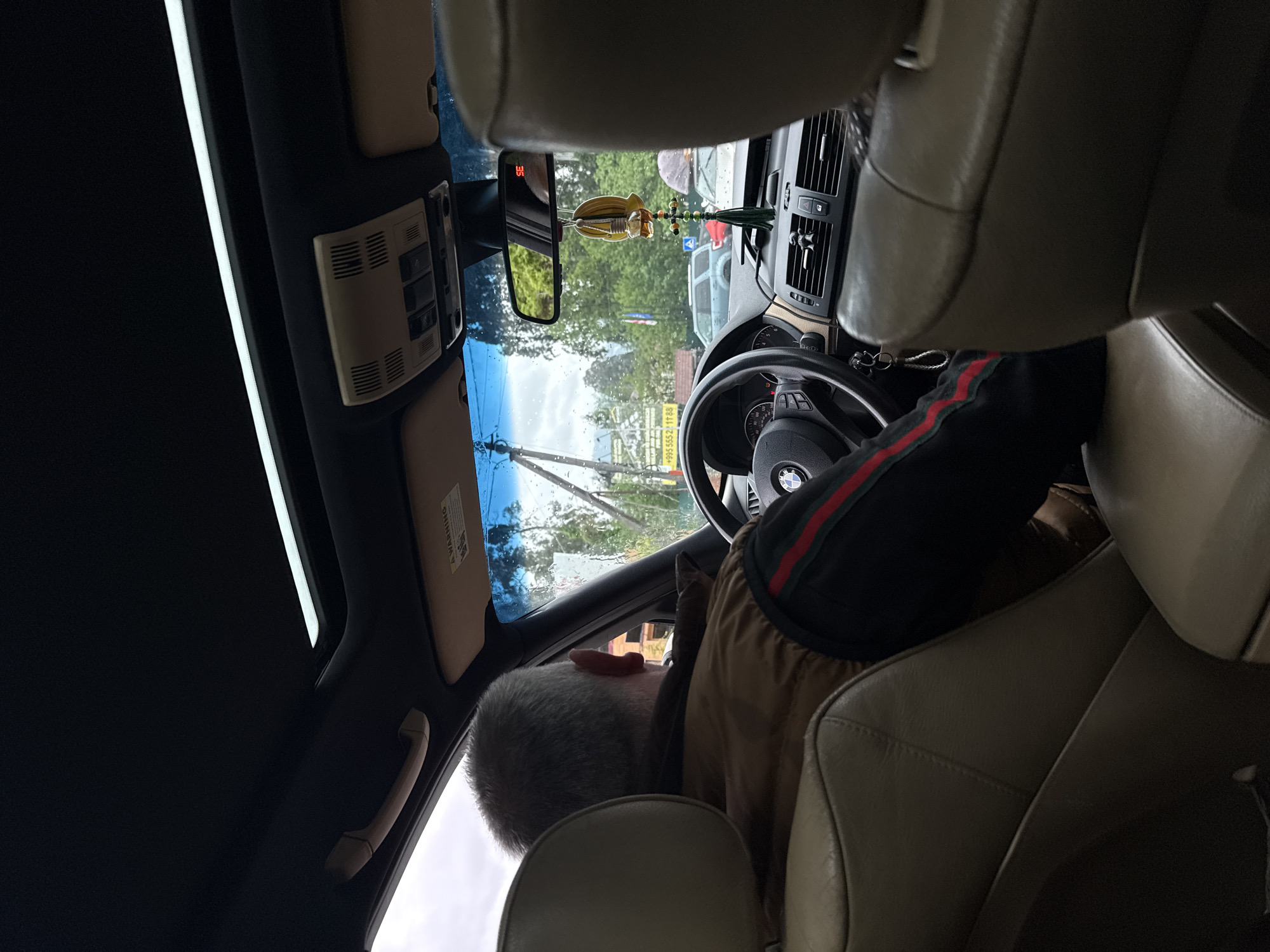 The photograph depicts the interior of a car, likely a BMW based on the steering wheel emblem, in what appears to be Chakvi, Georgia, as indicated by the visible telephone number "+995 555 211 88" on a sign outside. An older man with gray hair is in the driver's seat, wearing a tan jacket with a black sleeve bearing a red stripe, presumably getting ready to drive. The car has a sunroof and beige leather interior. A decorative tassel hangs from the rearview mirror. Through the windshield, the scene includes trees, power lines, and other cars, suggesting a residential or commercial area. Raindrops are visible on the windshield.