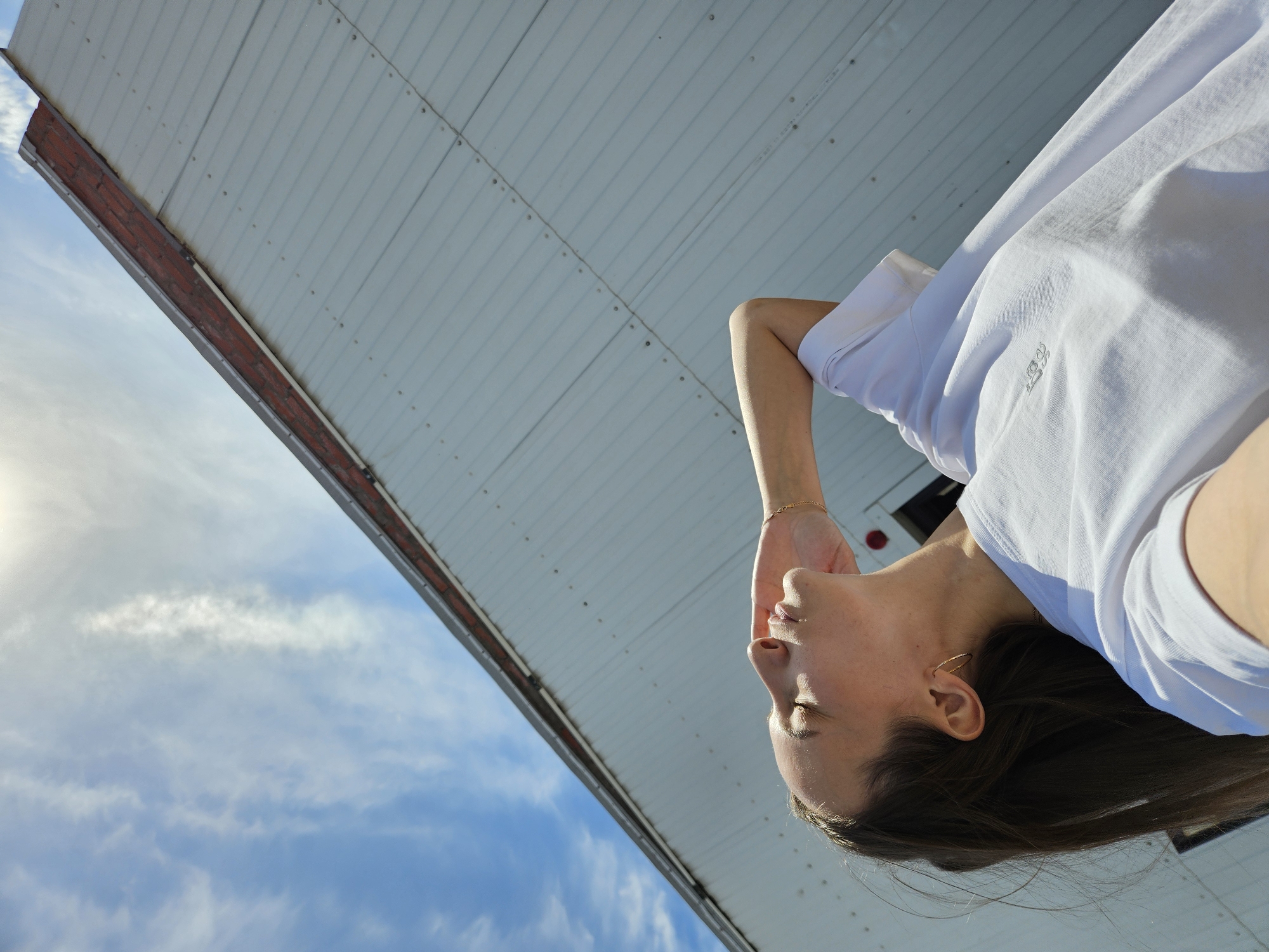 The image is an upward-angled view of a person standing outside, seemingly tilted backward, with the sky visible behind them. The person is wearing a white t-shirt and has brown hair, with a gold bracelet on one wrist and a gold hoop earring on one ear. Behind the person is a building's exterior wall or roof, predominantly white with linear patterns that suggest panels or siding. A brick element is also visible along the edge. The sky is blue with some clouds. The sun is visible on the left, with soft, diffused light.
No specific details in the image confirm that it's taken in the city of Chaikovsky, Russia.
