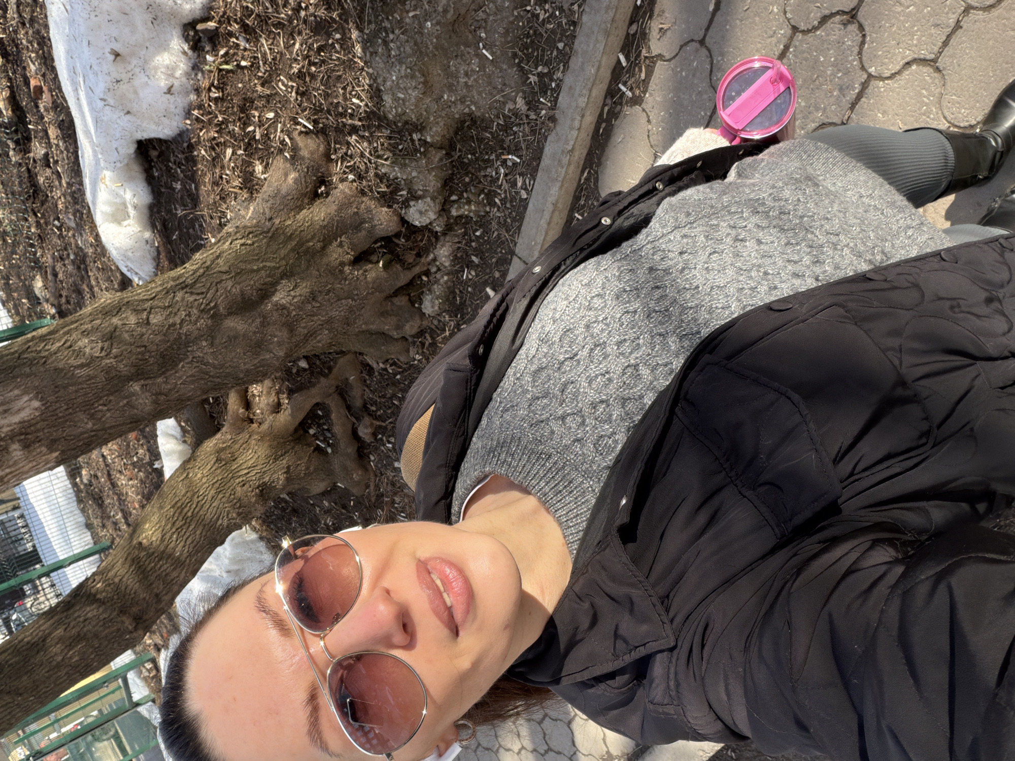 A woman wearing sunglasses, a grey sweater, a black jacket, grey leggings, and black boots is taking a selfie outdoors. She stands on a paved walkway beside a patch of dirt and snow, with a tree branch visible in the background. There is a pink cup with a straw next to her. The location is Perm, Russia.