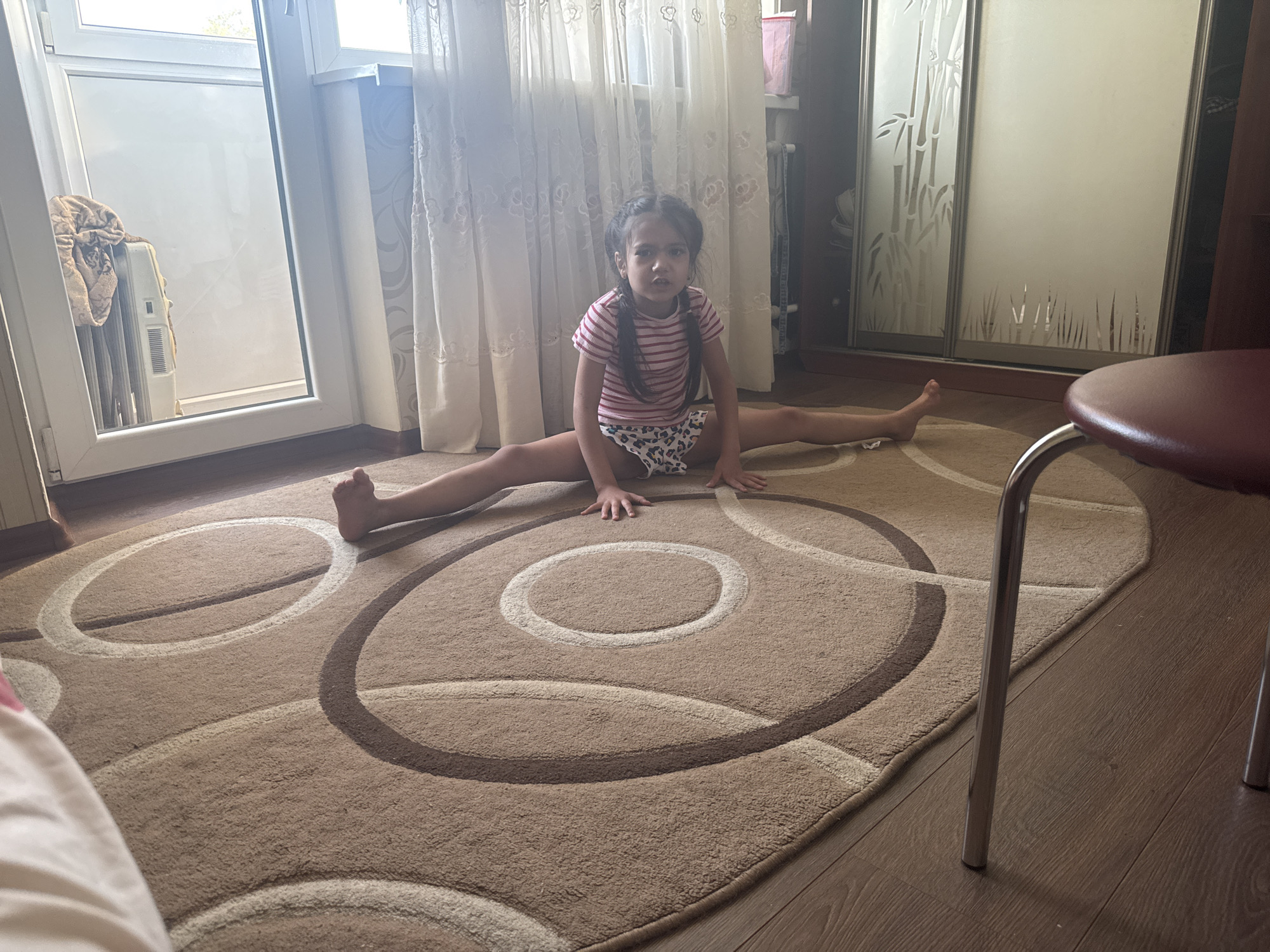 An indoor scene depicts a young female child performing a full frontal split on a patterned rug. The child, with dark hair styled in two pigtails, is wearing a pink and white striped short-sleeved top and white patterned underwear. Her hands are flat on the rug in front of her, and she looks directly at the camera.
The rug, which covers a significant portion of the wooden floor, is light brown and features a design of concentric and overlapping circles in varying shades of brown and white.
To the left, a large white-framed window or balcony door allows daylight into the room. Through the glass, a white radiator or appliance draped with a beige blanket or towel is visible, with a blurred outdoor background.
In the background, light-colored, translucent curtains with an intricate pattern hang across a wall. Behind the right portion of the curtains, a section of wall with a subtle patterned wallpaper is discernible.
To the right, a multi-paneled sliding door closet or wardrobe is present. The left two panels feature a frosted or etched glass design depicting bamboo stalks and grasses. The rightmost visible panel is solid and light-colored. A dark brown wooden frame of the closet or another furniture piece is visible on the far right, with a glimpse of stored items.
The lower portion of a chair with a dark red seat and a chrome-plated metal leg is partially visible in the bottom right corner of the frame. A pink container is visible in the upper right background, above the closet. The location is in the city of Unknown, Unknown.