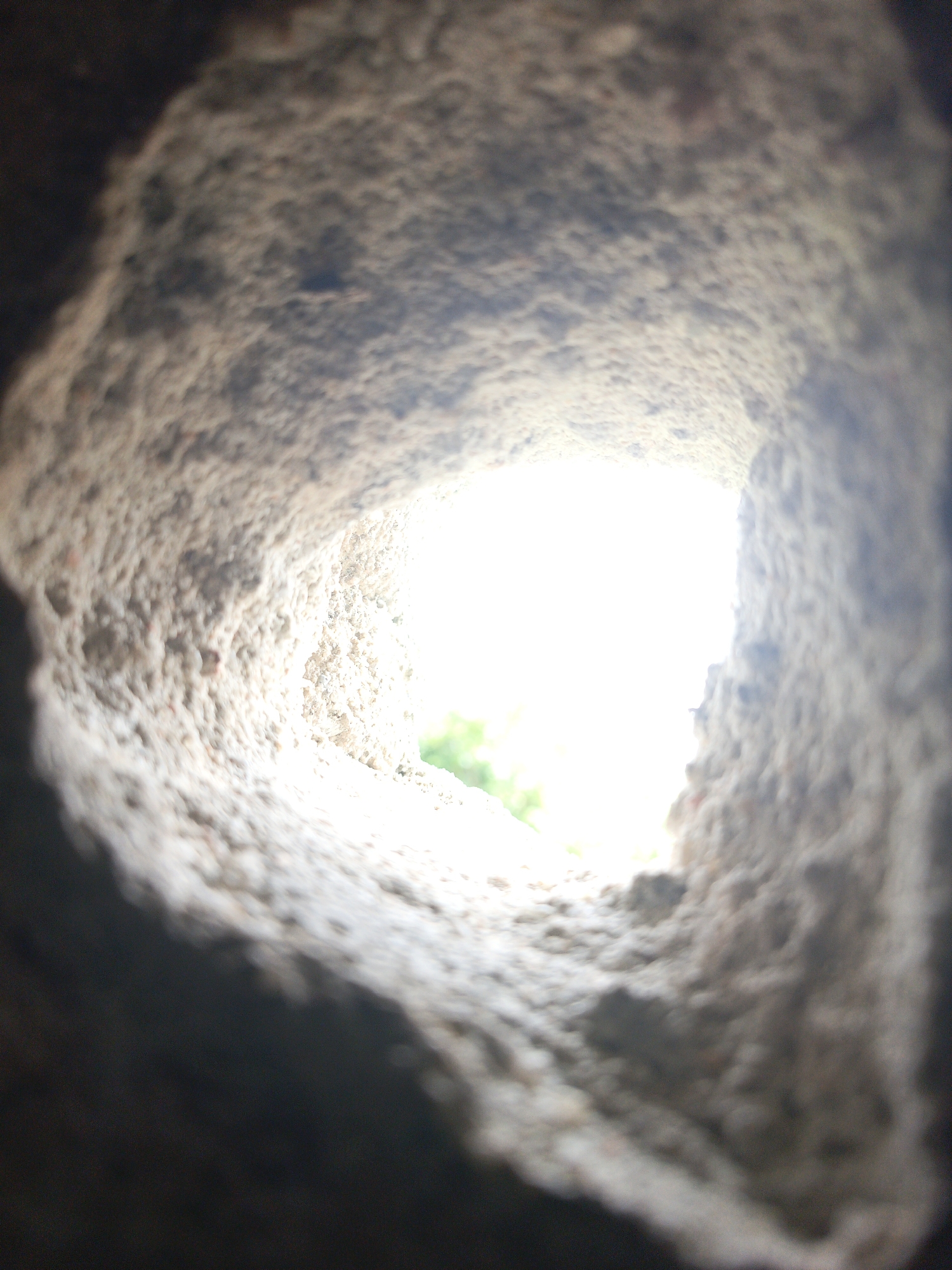 The image shows a close-up view looking through a rough-edged hole, likely in a concrete or stone structure. The interior of the hole is textured and pale, suggesting the material's composition. Outside the hole, a bright light source is visible, with a hint of green foliage indicating an outdoor environment. The location is Unknown, Unknown.
