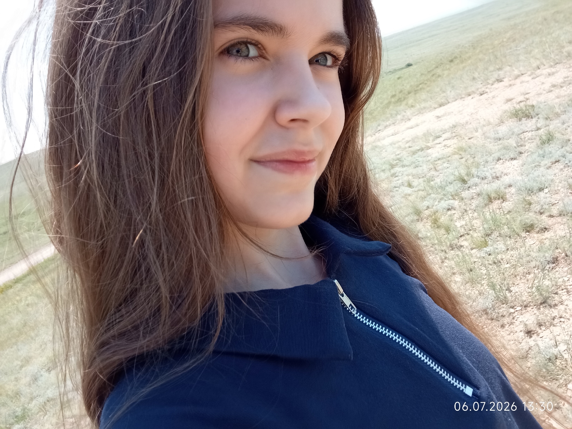 A medium shot shows a young woman with long, brown hair, looking to her right with a slight smile. She is wearing a dark blue zippered top. The background is a grassy, undulating landscape, likely a field or steppe. The image has a timestamp in the lower right corner reading "06.07.2026 13:30". The location is identified as Aleksandrovka, Kazakhstan.