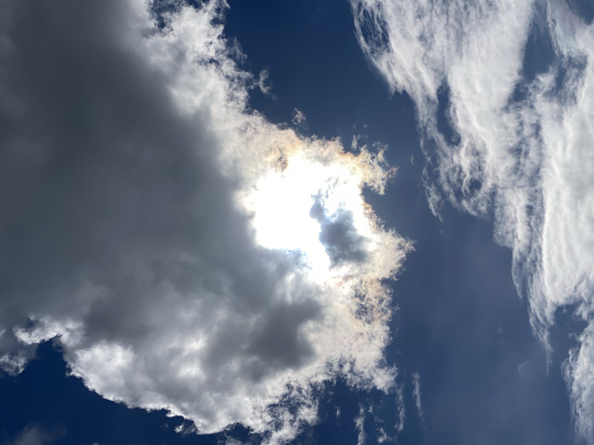 The image depicts a daytime sky in Ustyvytsia, Ukraine. The sky is predominantly blue, with a few scattered white, wispy cirrus clouds visible on the right side of the image. A large, gray cumulonimbus cloud is present on the left side, partially obscuring the sun. The sun is shining brightly through a gap in the clouds, creating a halo effect with tinges of brown/orange surrounding the edges of the clouds nearest the sun. No people, buildings, or other objects are visible in the picture.