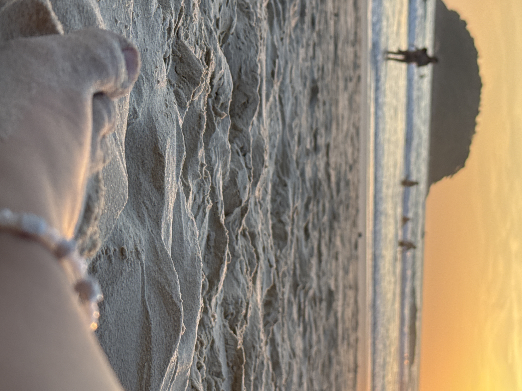 A low-angle, close-up shot captures a person's sandy foot and ankle in the foreground, adorned with a bracelet of small, light-colored beads. The background is blurred, depicting a beach scene at sunset in Karon, Thailand. Gentle waves are visible lapping onto the shore, and several indistinct figures of people are present in the water, some wading and others appearing to be swimming or surfing. A dark, treed island or large rock formation is silhouetted against the warm, orange glow of the setting sun, which dominates the sky. The sand in the foreground displays rippled textures from the receding tide.