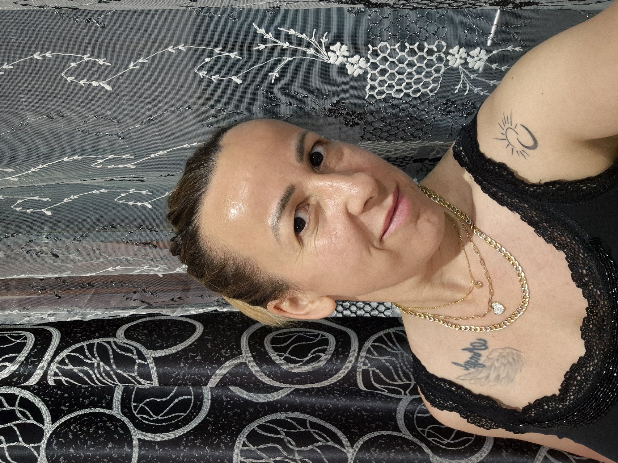 A woman with blonde hair pulled back in a ponytail, wearing a black lace top and several gold necklaces, smiles at the camera. She has a tattoo of a wing and text on her chest, and a tattoo of a crescent moon and sun on her right shoulder. She is standing in front of a patterned curtain.
