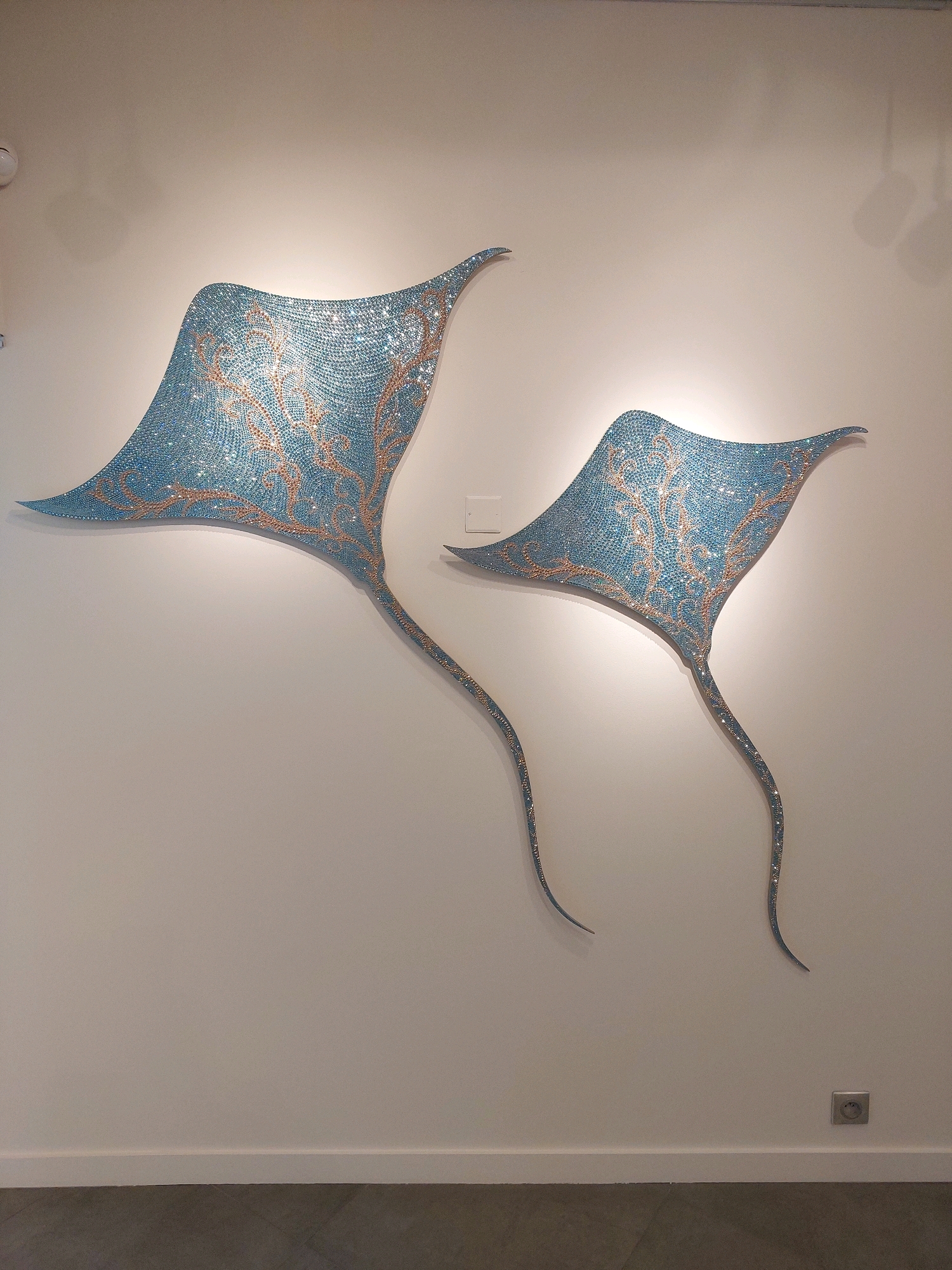 The image shows two decorative stingray sculptures mounted on a white wall in what appears to be an art gallery or exhibition space. The sculptures are made of small, iridescent blue tesserae and feature swirling golden patterns. The larger stingray is on the left, and a smaller one is to its right. Both sculptures have long, trailing tails. The wall is lit by spotlights, casting subtle shadows. A white baseboard runs along the bottom of the wall, and a dark floor is visible below. A light switch and an electrical outlet are present on the wall. Based on the context of Saint-Paul-de-Vence, this scene is likely from the Fondation Maeght, a renowned modern and contemporary art museum.