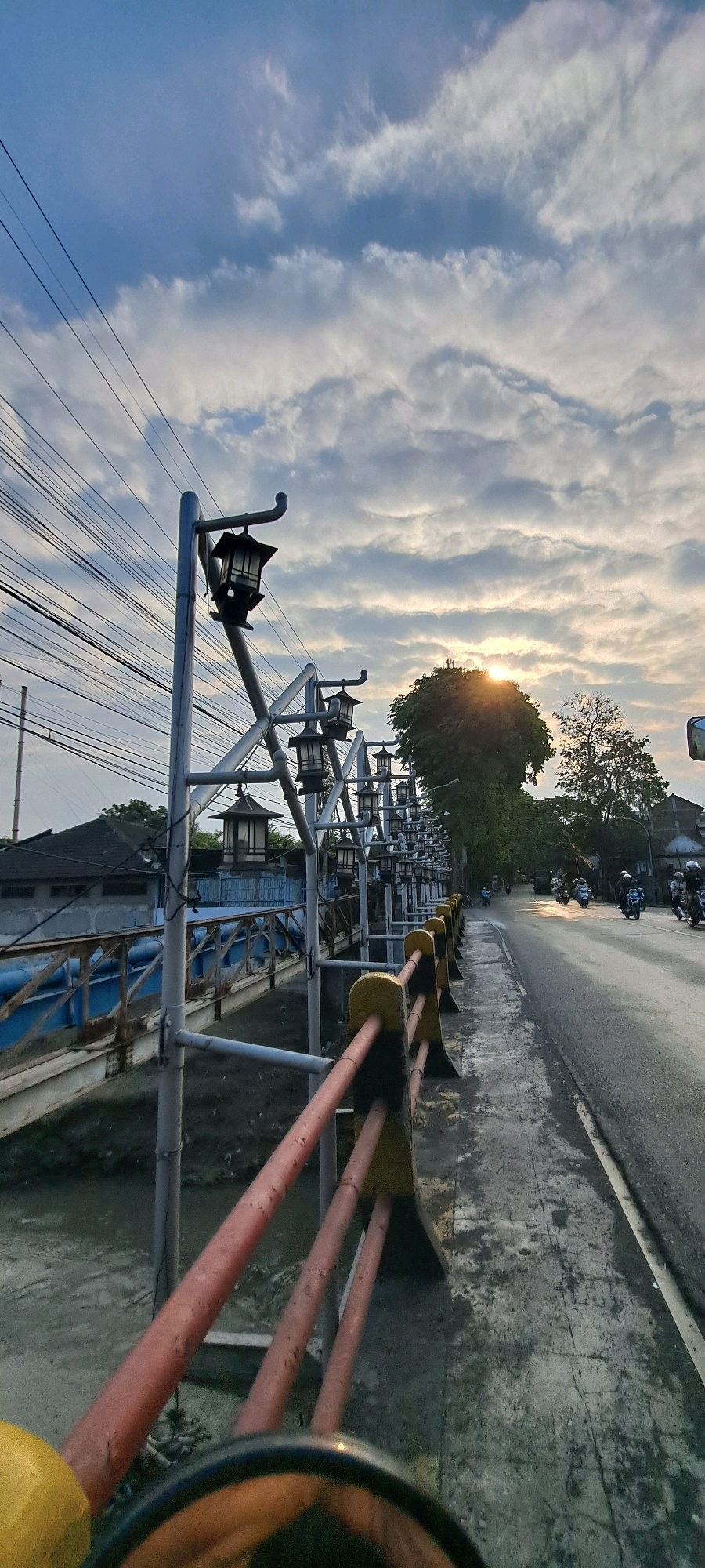 The image depicts a street scene in Pasuruan, Indonesia, during what appears to be sunset. The foreground features a yellow and red metal railing alongside a grey concrete sidewalk. To the left, a bridge is visible with blue railings and a structure resembling a building. A series of decorative lampposts with dark, oriental-style shades line the sidewalk. The background shows a road with several motorcycles and their riders. Lush green trees and some buildings are present on the right side of the road. The sky is filled with a mix of white and grey clouds, with the sun partially obscured behind a large tree, casting a warm glow. Power lines are prominently visible crisscrossing the upper left portion of the frame. The overall impression is of an urban environment transitioning into evening.