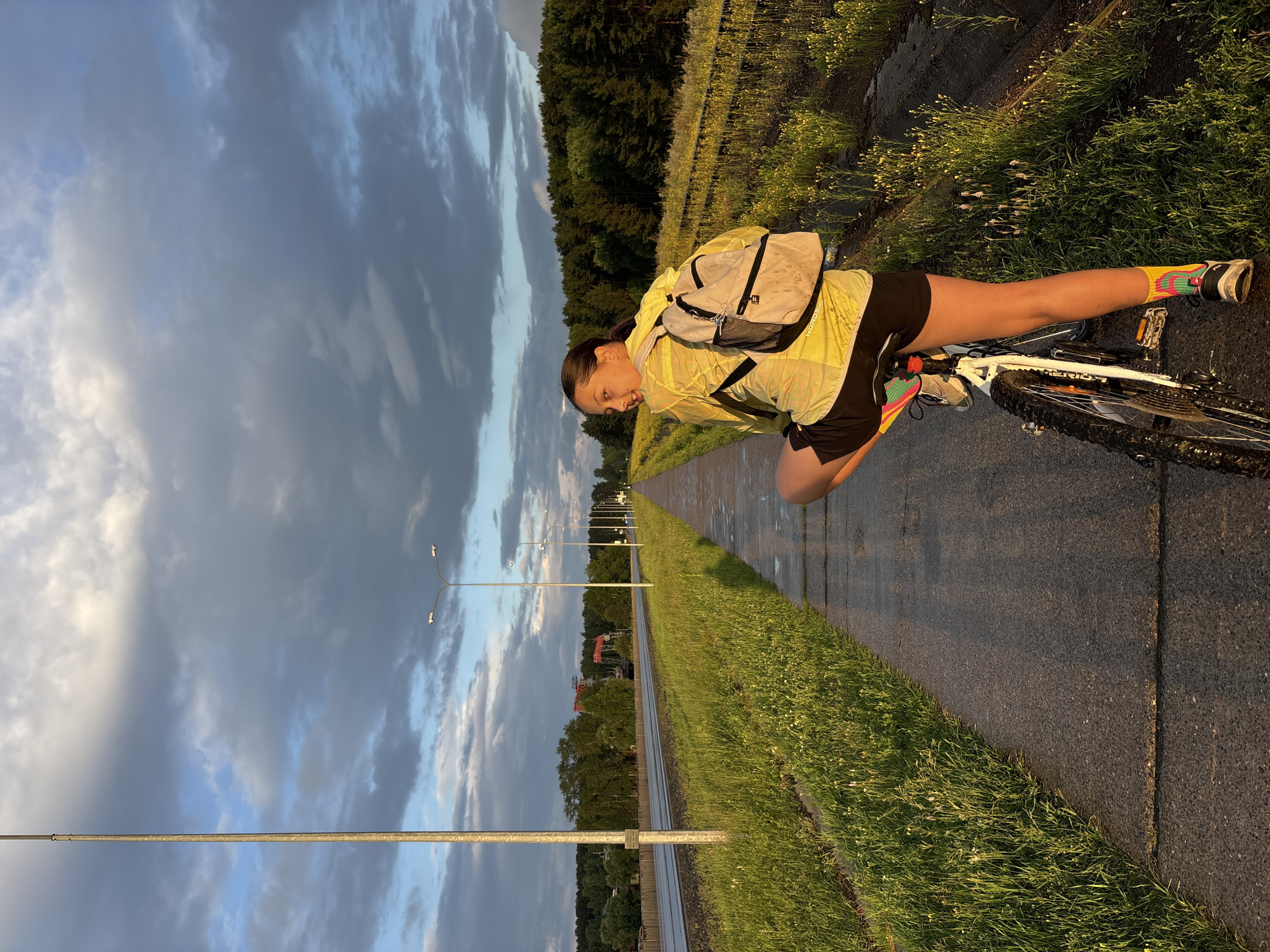 A young woman with dark hair pulled back, wearing a bright yellow jacket, black shorts, and colorful socks, is posed on a white bicycle on a paved path. She is looking back over her shoulder with a smile. The path runs alongside a grassy area with wildflowers and a road with streetlights. In the background, a dense forest lines the horizon, and a cloudy sky with dramatic lighting suggests either sunrise or sunset. The scene appears to be on the outskirts of Chaikovsky, Russia, possibly near a major road or a recreational path.