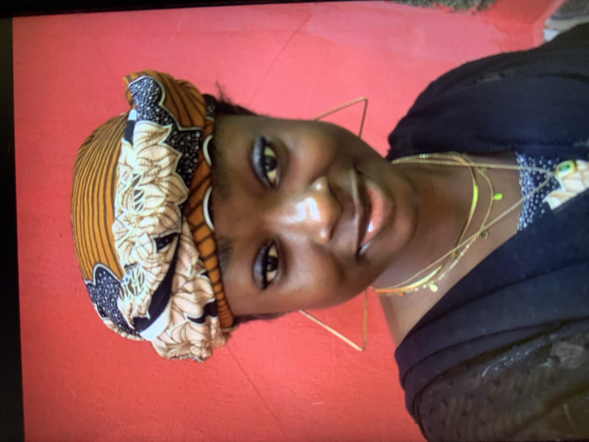 The image shows a woman with dark skin wearing a patterned headwrap with orange, brown, and black accents. She is smiling and has layered gold necklaces. She is also wearing gold square hoop earrings. The background is a solid red color. There is no evidence in the image to suggest this image was taken in Kabong, Nigeria, or any other specific location.