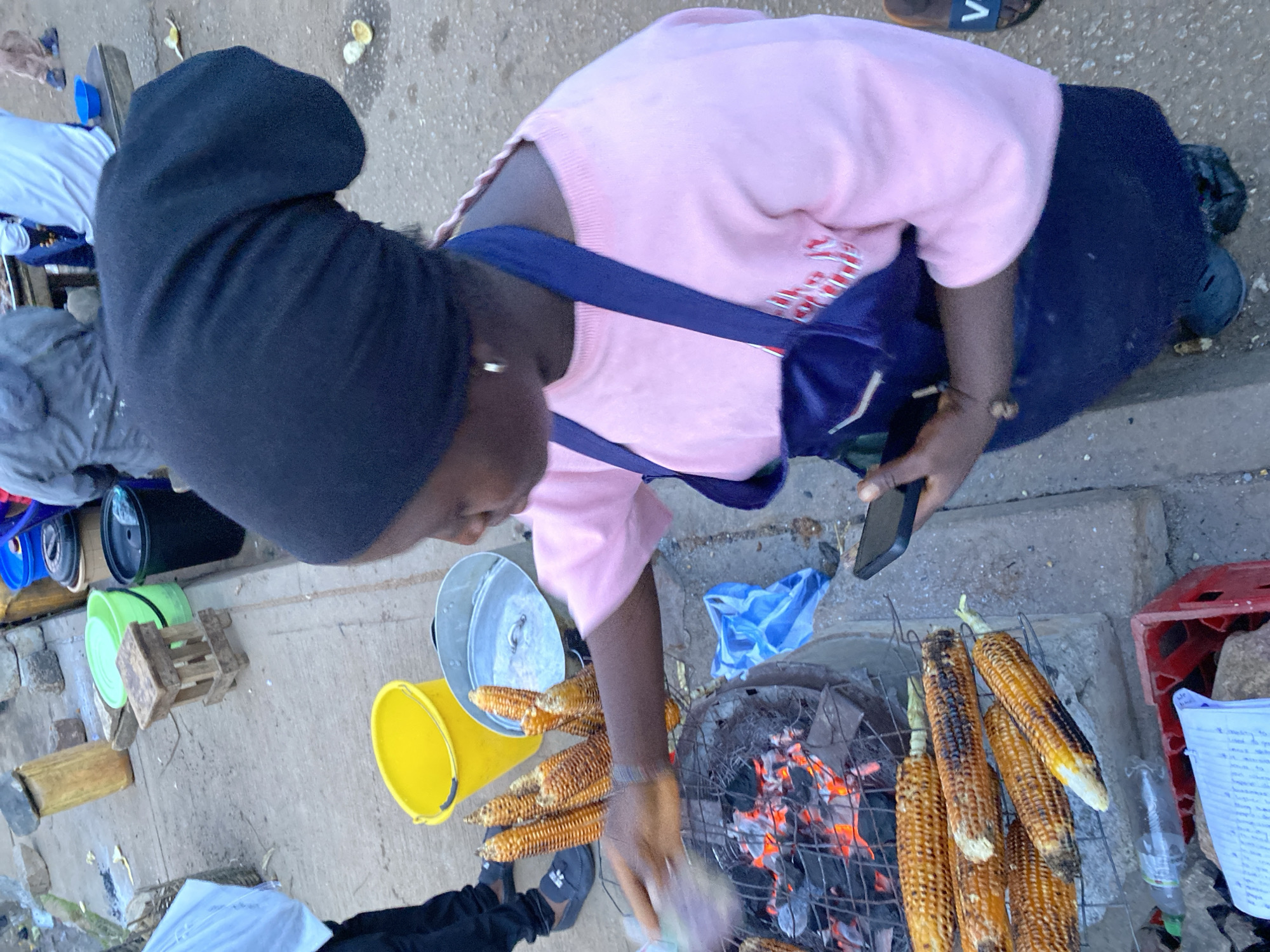 In Bukuru, Nigeria, a woman wearing a black headscarf and a pink shirt with a dark blue apron is preparing corn on a grill. The corn is arranged on a metal rack over hot coals. Several ears of corn are visible, some appearing to be partially cooked or roasted. In the background, a yellow bucket, a metal pot, and other assorted items are present. A blue plastic bag lies on the ground near the grill. The woman is holding a smartphone in her right hand. The ground appears to be paved with concrete and dirt.
