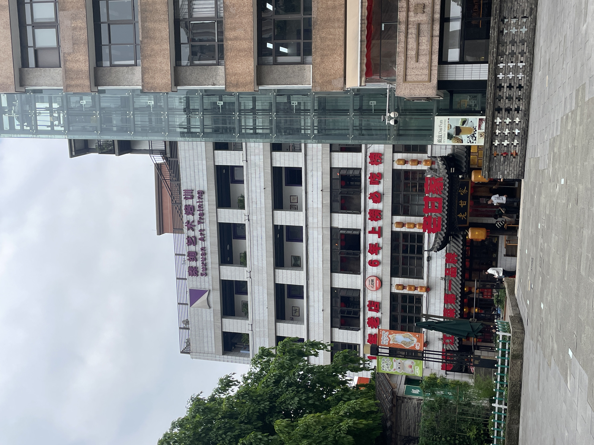 The image captures a street scene in Yuzhong District, China. Prominent in the frame is a tall, multi-story building with white tiled walls and dark-framed windows. A glass-enclosed skybridge connects this building to another structure, distinguished by its brown exterior and similar windows. Red Chinese characters are prominently displayed on the side of the white building. Below, a restaurant area with traditional Chinese architectural elements is visible. A sign in Chinese reads "Lao Gan Jia" (老甘家), and an advertisement for a coffee shop, "Dai Fun," can be seen. The sign on top says "Su Xuan Art Training".
The foreground features foliage from trees and other greenery. A paved sidewalk extends into the frame, suggesting an urban environment. A person is partially visible near the restaurant entrance. The sky is overcast, giving the scene a muted, gray tone.