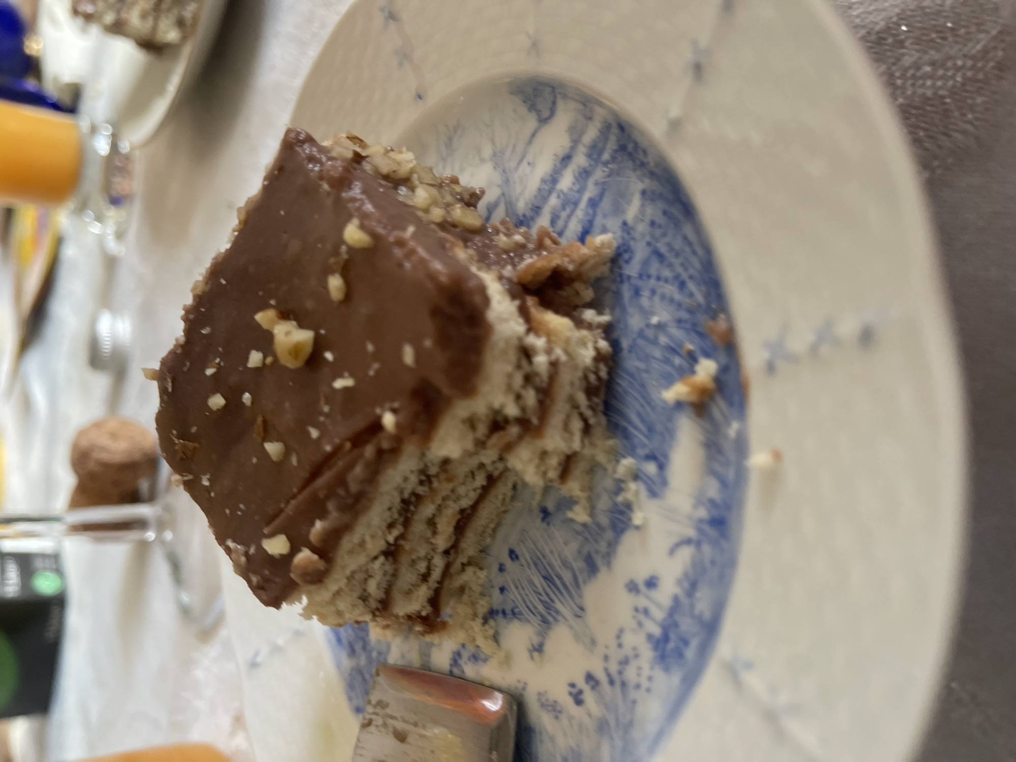 A high-angle close-up shot shows a piece of layered chocolate cake on a blue and white patterned plate. The cake is rectangular and has a dark chocolate topping sprinkled with chopped nuts. The layers beneath the chocolate appear to be composed of a light-colored cream or frosting alternating with what might be biscuit or cake layers. A metal utensil, likely a knife or serving spatula, is partially visible underneath the cake. The plate rests on a table, and other items are visible in the background, including a glass of orange liquid, another dessert, and a small container. The scene appears to be indoors and is likely a domestic setting. The location in Maidstone, United Kingdom is not determinable from the image.
