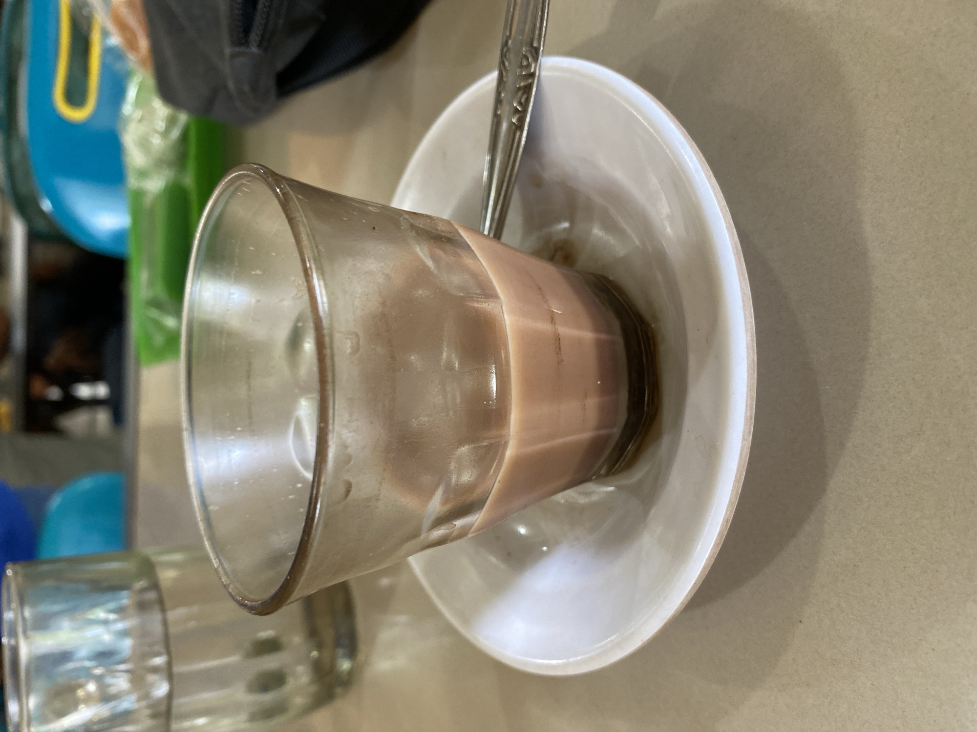 The image shows a close-up of a glass filled with a light brown liquid, possibly coffee or chocolate milk, resting on a white saucer. A silver spoon is partially submerged in the liquid. The glass is transparent, and some condensation is visible on its exterior. The saucer is round and white, with a slightly raised rim. The background is blurred, but suggests an indoor setting with a textured surface, likely a table, and hints of other objects like a blue and green plastic container and a dark bag or backpack. The lighting appears to be artificial, casting a shadow from the glass onto the saucer. The overall impression is a casual, possibly breakfast or afternoon break scene, in a domestic or cafe environment. There is no visible text in the image.