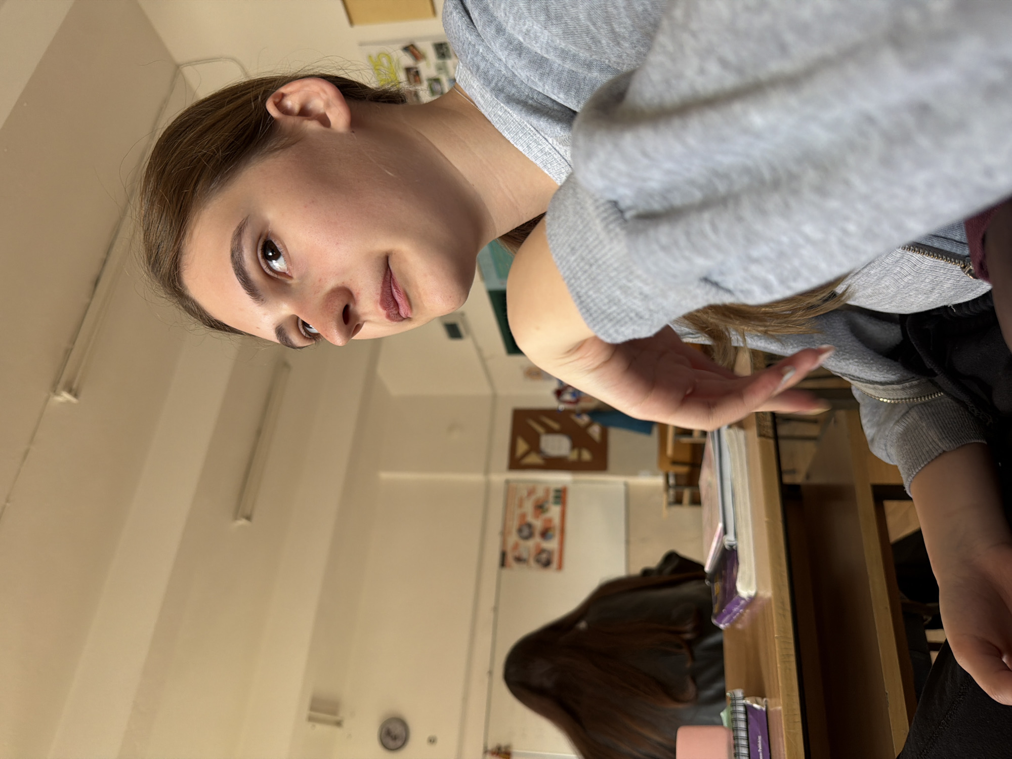 A young woman is captured in a close-up, slightly upward-angled shot in what appears to be a classroom or study environment in Panagyurishte, Bulgaria. She is positioned on the right side of the frame, looking towards the camera with a neutral to slightly inquisitive expression. Her brown hair is pulled back, and she is wearing a light gray hooded sweatshirt with a silver chain bracelet visible on her left wrist. Her left hand is raised slightly, palm open, as if in a gesture or reaching out.
In the background, the long, dark hair of another person is visible, seated at a wooden desk in the mid-ground, facing away from the camera. The desk holds several notebooks or books, some with purple covers and one pink item. On the light-colored walls in the background, several educational posters or charts are visible, though their specific content is largely indistinct. One appears to show diagrams or illustrations. Fluorescent light fixtures run along the ceiling, and a round wall clock is visible on the far wall. The overall setting suggests an institutional or educational building. On the spine of one of the purple notebooks, the word "Publishing" is partially legible.