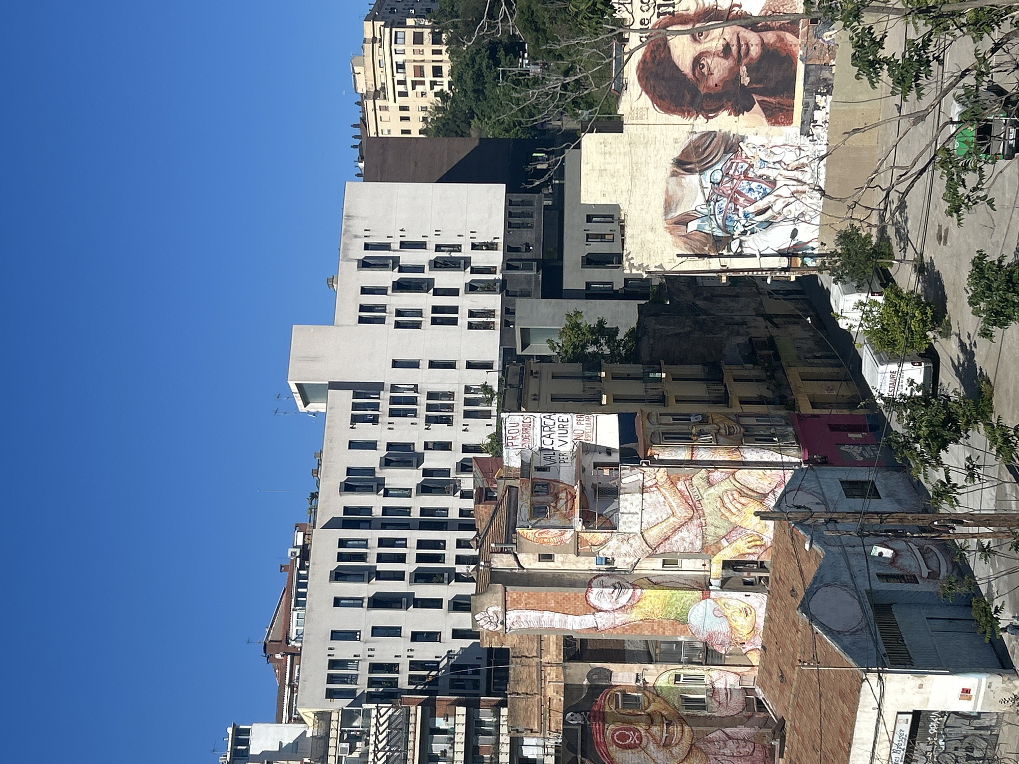 This media file captures a vibrant, sun-drenched urban street scene in Barcelona, showcasing a blend of architectural styles and prominent street art. The sky above is a clear, brilliant blue, indicating fair weather during what appears to be mid-morning or early afternoon, with strong sunlight casting crisp shadows.
The scene is dominated by a collection of buildings. On the left, a tall, modern structure with a light-colored facade features numerous rectangular windows. Adjacent and throughout the mid-ground are older buildings, some with brickwork and others painted, which serve as canvases for extensive murals.
Several large-scale murals are key subjects. In the upper right, a large, expressive portrait of a person with long hair and a beard, rendered in warm, earthy tones, gazes upwards. Below this, another mural depicts abstract or figurative elements in blues and whites. Further down and to the left, multiple other murals on different facades display stylized human figures, often with elongated limbs, intricate patterns, and bright colors, including one with a distinct blue circle on a face.
Sparse green foliage from trees and some bare branches are scattered across the view, providing natural contrasts to the concrete environment. Visible text on a sign in the mid-ground reads "PROU D'IDERROCS" (Enough demolitions) and "VAL CÀRGA PER VIURE," indicating a social or political message. Other smaller signs, such as "RESTAURA," and graffiti like "EL BOKASER" are also present. The overall atmosphere suggests a lively, culturally rich urban area where art plays a significant role in defining the street's character.