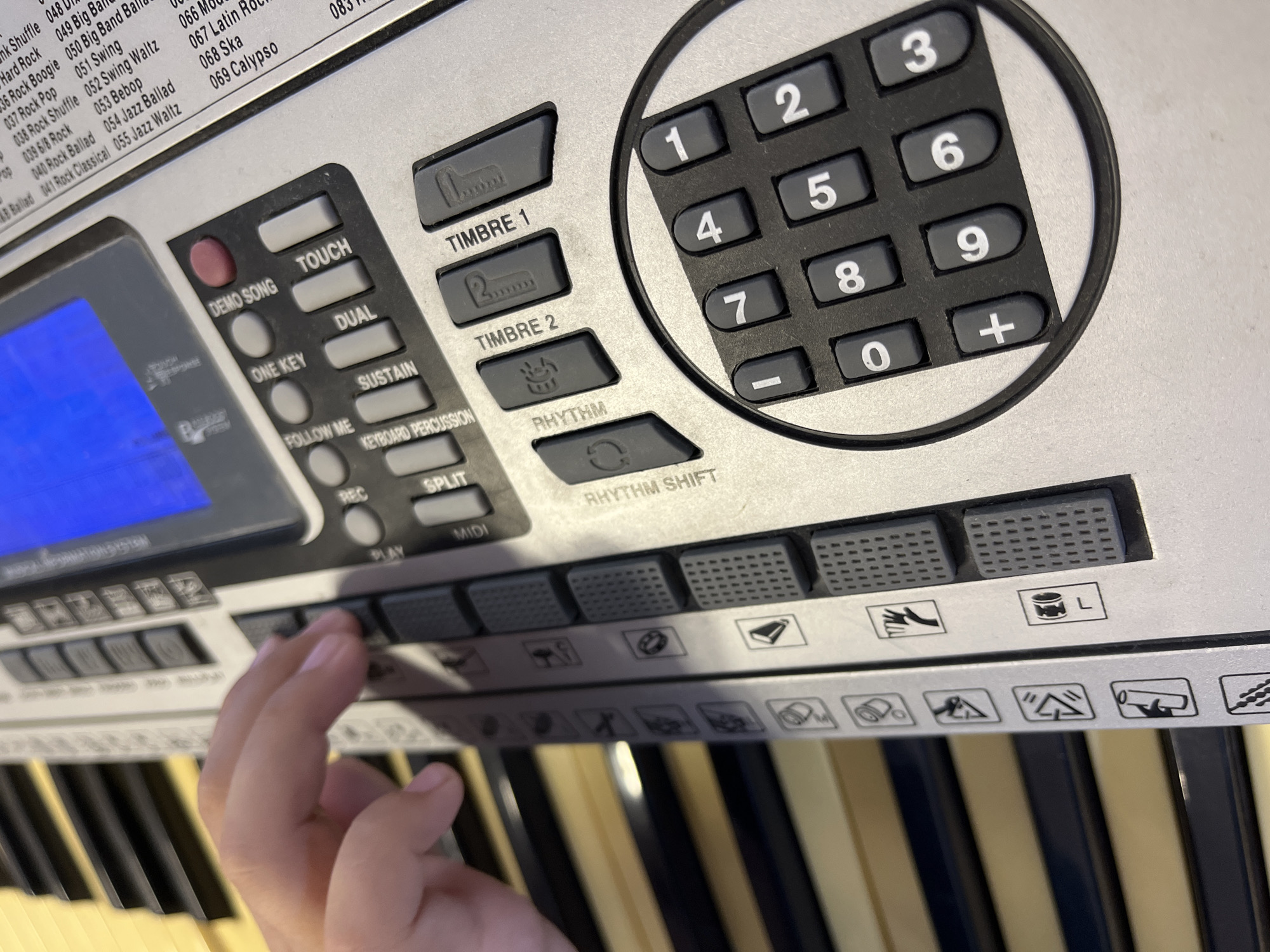 The image shows a close-up view of a digital keyboard with a child's left hand pressing one of the keys. The keyboard is silver and features various buttons, including a numeric keypad, "DEMO SONG", "ONE KEY", "DUAL", "TOUCH", "SUSTAIN", "RHYTHM", "RHYTHM SHIFT", "TIMBRE 1", and "TIMBRE 2". There are also icons and labels printed on the keyboard, some of which are partially visible. The background shows the black and white keys of the keyboard. The image is taken from a slightly elevated angle, focusing on the hand and the control panel of the keyboard. There is no visible indication of the city of Krasnyye Barrikady, Russia, in the image.