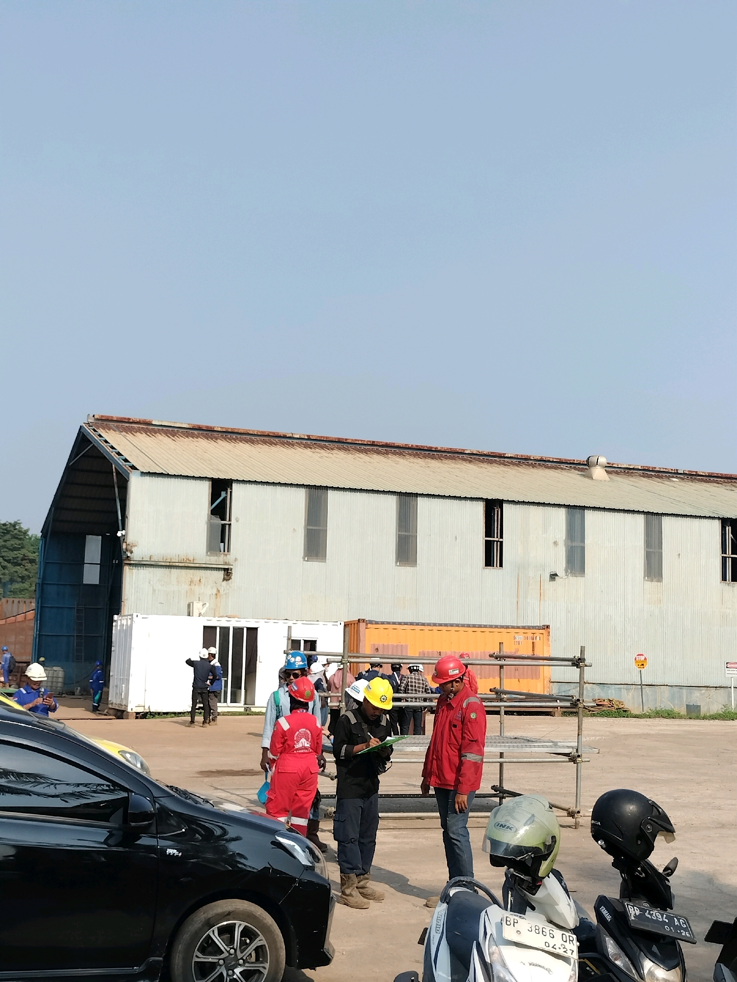 The image depicts an outdoor scene, likely a construction or industrial site, under a clear sky. In the background, a large, weathered, corrugated metal building stands with several windows. In front of this building, there are several modular structures and shipping containers.
In the foreground and midground, a group of individuals, mostly wearing hard hats and work attire, are gathered. Some are engaged in conversation or appearing to review documents. Two individuals in red jackets and helmets are prominent, one appears to be interacting with a clipboard. Several motorcycles are parked in the immediate foreground.
The setting appears to be an open, paved area, possibly a yard or lot, with some scaffolding visible. The overall impression is one of an active work environment. Based on context clues in the cropped images, such as license plates with "BP" which is common for Batam, Indonesia, and the type of industrial structures visible, this scene is likely located in Batam, Indonesia.