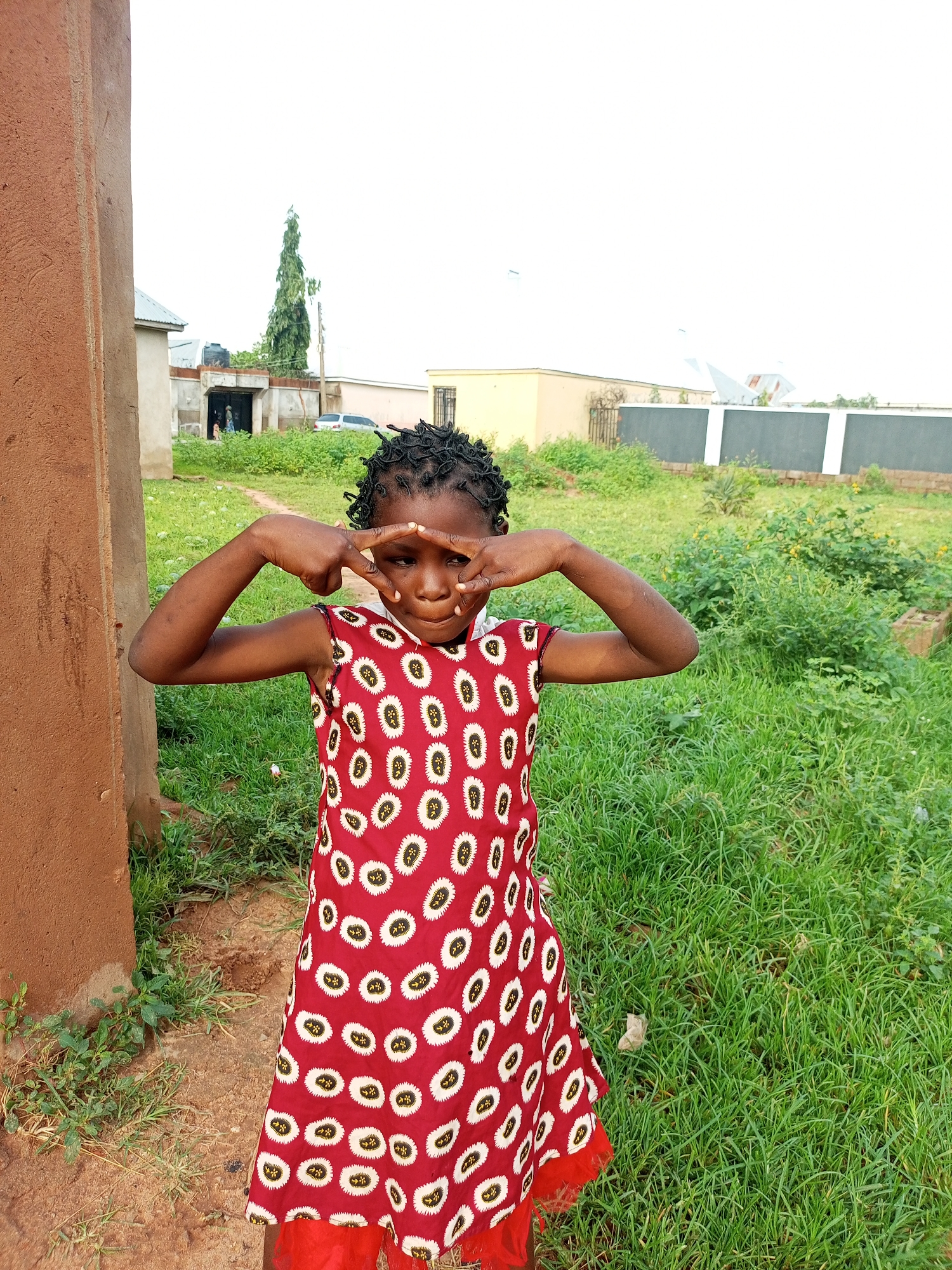 A young Black girl with braided hair stands in front of a brown stucco wall, looking at the camera with her hands held up to her eyes, forming a frame. She is wearing a sleeveless red dress with a white and black oval pattern. Behind her, a grassy yard stretches towards a fence and buildings in the distance. The scene appears to be in Gombe, Nigeria.