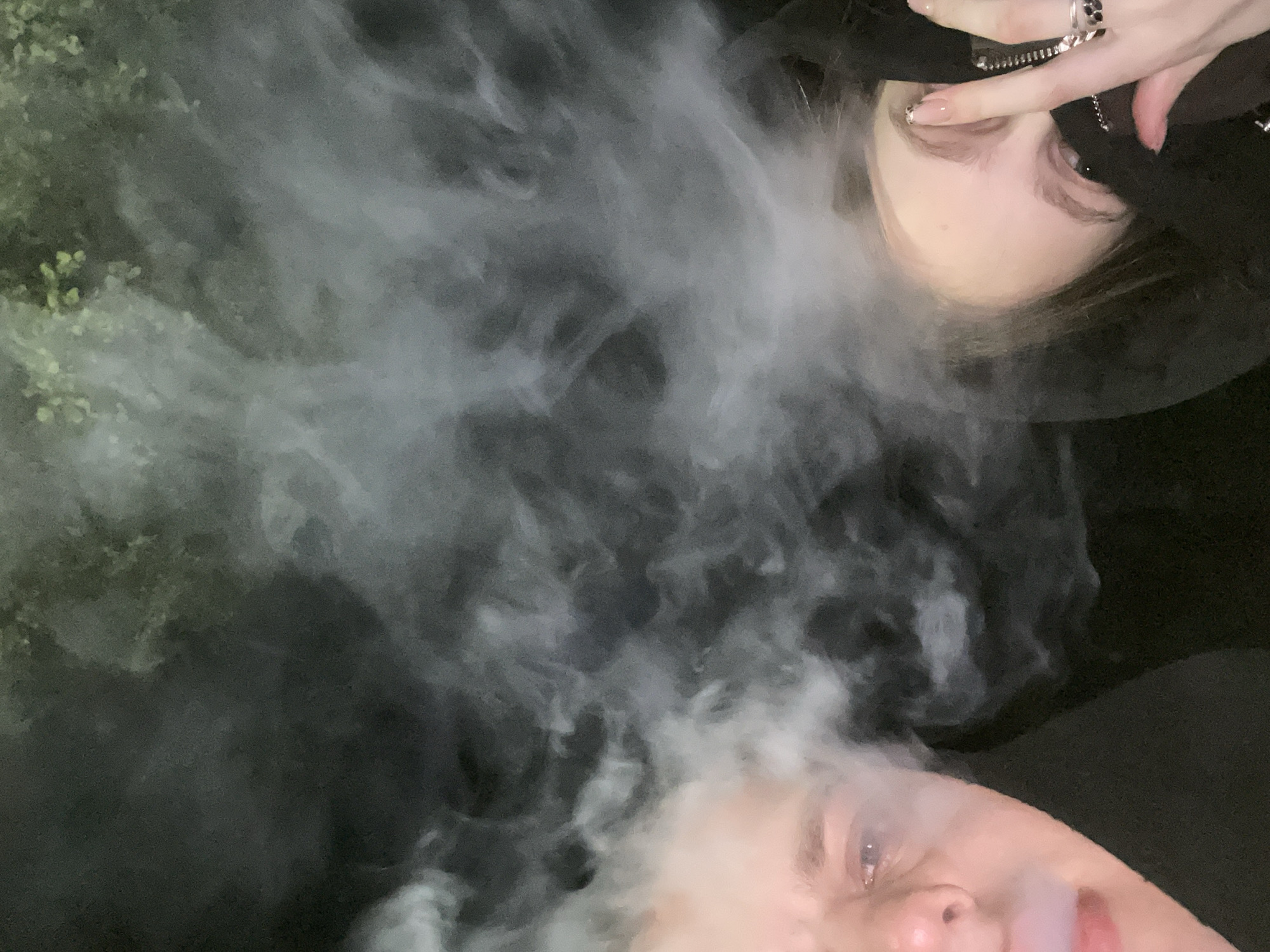 A vertically oriented, low-light image captures two individuals partially visible, separated by a dense cloud of white vapor. In the upper-right corner, the eye, nose, and forehead of one person are visible, with fair skin and dark hair. Their right hand, adorned with several rings (one with black stones, another silver) and manicured nails featuring French tips with black accents, is raised near their face.
In the lower-left corner, the eye, nose, and mouth of a second person are visible, featuring fair skin and light hair. This individual is actively exhaling a significant plume of white vapor or smoke, which billows from their mouth and spreads across the central and left portions of the frame. A dark, leafy green plant is partially visible in the upper-left corner, some of its foliage obscured by the vapor.
The overall background is dark, suggesting a nighttime setting or a dimly lit indoor space. The substantial amount of vapor implies the use of a vaping device. No text is visible in the image. The scene is located in Красные Баки, Russia.