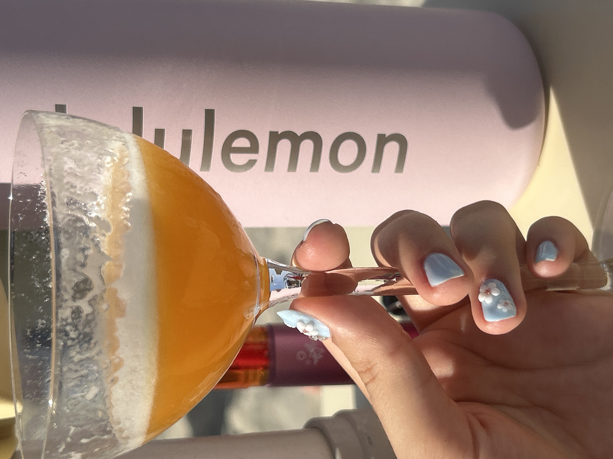 A person's hand, with light blue nail polish and small floral decorations on one nail, is holding a stemmed glass filled with an orange beverage. The beverage has a frothy, layered appearance, with white foam on top and orange liquid below. The glass is positioned in front of a pale pink object with the word "lululemon" printed in silver letters. The setting appears to be outdoors, with sunlight casting shadows, suggesting it is daytime. The overall mood is bright and cheerful. The background includes hints of other objects, including a dark purple bottle. The location context is Cupra Marittima, Italy.