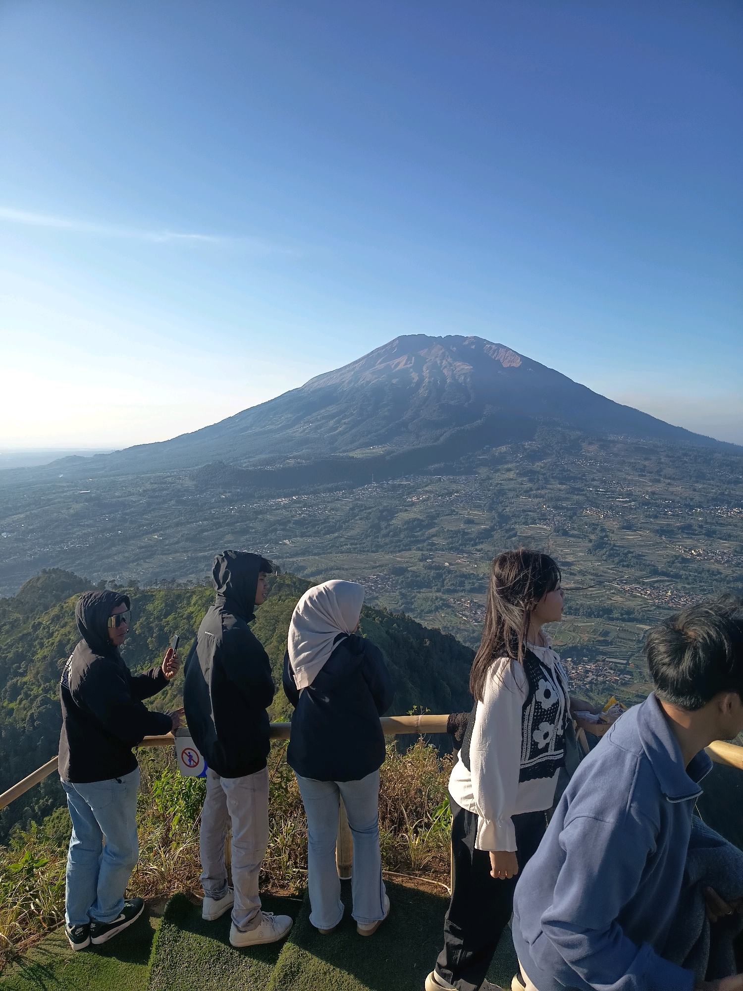 In Keditan, Indonesia, five individuals are positioned on a raised viewpoint overlooking a valley and a large mountain. The viewpoint features a wooden railing and a green, artificial turf surface. From left to right, the first person is a male wearing sunglasses, a black hooded jacket, and light blue jeans, holding a phone in his right hand. Next is another male in a black hooded jacket and light gray pants. To his right, a female wears a light-colored hijab and a dark hooded jacket with light blue jeans. Further right, a young female with long dark hair sports a white blouse with black floral embroidery over dark pants. The furthest individual on the right, partially visible, is a male in a blue zippered jacket. The background reveals a vast, verdant landscape with a prominent mountain, likely Mount Merapi, under a clear blue sky. The valley below is dotted with scattered buildings and agricultural fields, suggesting a rural or semi-rural setting. The time of day appears to be morning or late afternoon, given the lighting and long shadows.