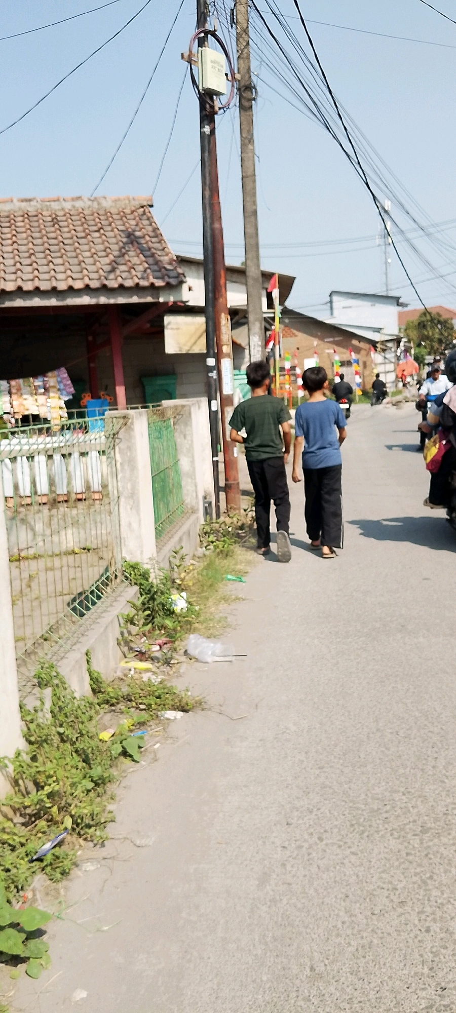 The image shows a street scene in Karangrahayu, Indonesia. Two young males are walking away from the camera down the center of a paved street. The male on the left wears a dark green t-shirt and black pants, while the male on the right wears a blue t-shirt and black pants. To the left of the street is a fenced area with a building behind it, possibly a shop, with goods displayed outside. Overgrown vegetation and scattered trash are visible along the side of the road and in the fenced area. A utility pole with multiple wires is positioned on the left side of the street, and another pole is visible further down the street. The sky is clear and blue. In the background, more buildings and utility poles are present, along with several motorcycles. Red and white flags are visible along the street, suggesting a local celebration or event.