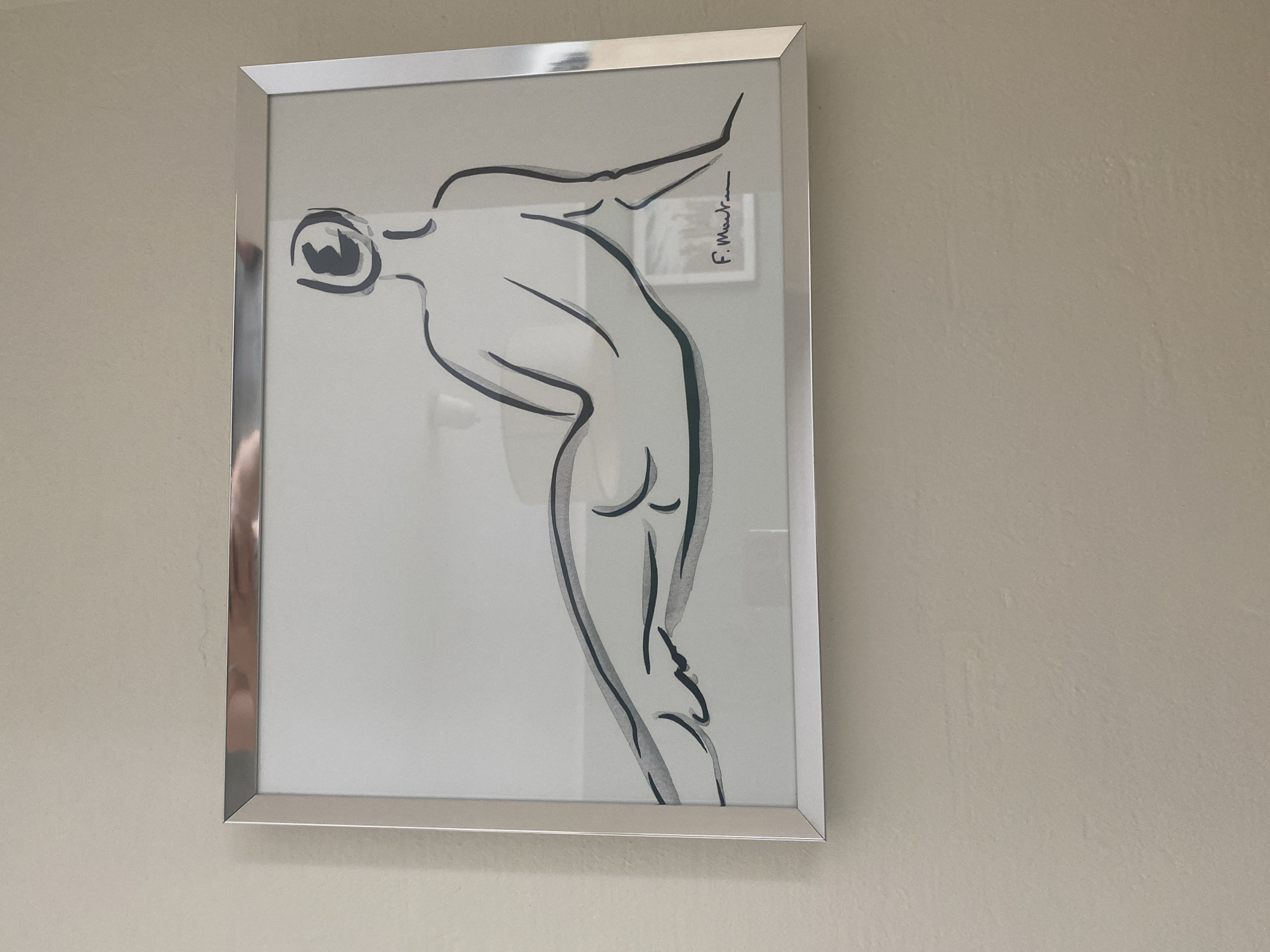 A framed artwork of a reclining nude is displayed on a neutral-colored wall. The artwork is a line drawing with watercolor accents, depicting a figure from behind with their head turned. The frame is silver and thin. The signature "F. Mauran" is visible in the lower right corner of the artwork. The scene is indoors, with the wall appearing to be a typical interior wall. There are no other discernible objects or people in the immediate vicinity of the artwork.