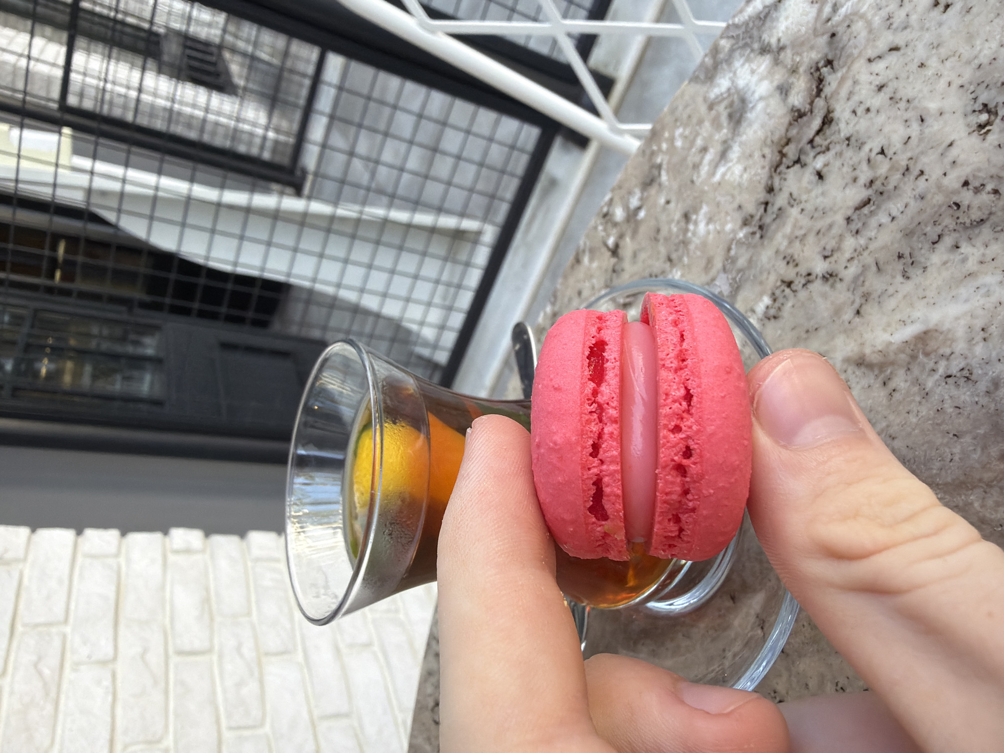 A close-up, over-the-shoulder shot shows a hand holding a pink French macaron above a glass of amber-colored tea with a lemon slice. The macaron has a creamy pink filling. The tea is in a small, clear glass with a saucer, sitting on a speckled granite tabletop. In the background, a white brick wall is visible to the left, and to the right, a metal grate partially obscures a white curtain or blind. The scene appears to be outdoors, possibly on a patio or balcony, as suggested by the lighting and the outdoor elements in the background. The location is likely Kemer, Turkey, given the context of the request.