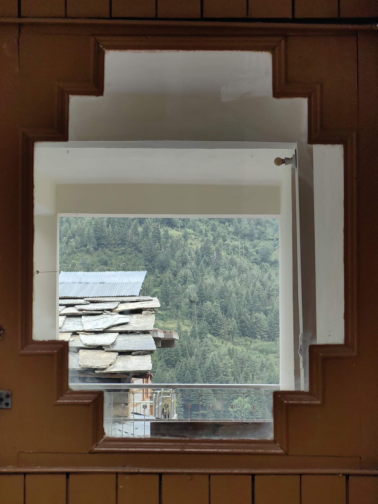 The image shows a view through a uniquely shaped wooden doorway into an outdoor scene in Vashisht, India. The doorway has a stepped, dark brown frame with a white interior. Through the opening, a dense forest of evergreen trees covers a hillside. In the foreground of the outdoor scene, there is a structure with a corrugated metal roof and layered stone roofing. A metal railing is visible below the stone structure. The lighting suggests it is daytime, possibly overcast, with the natural light illuminating the outdoor landscape. There are no people visible in the image.