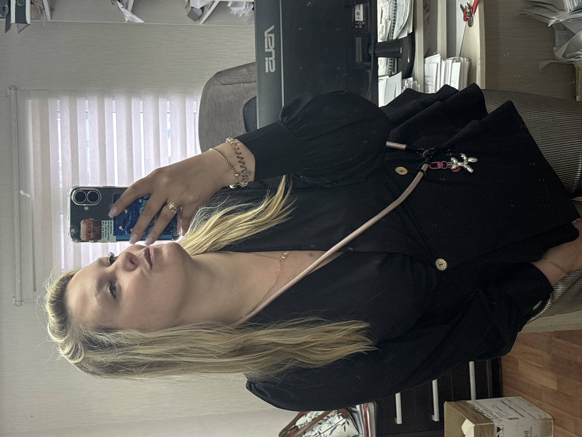 A woman is taking a selfie in what appears to be an office setting. She is holding a smartphone with a clear case and a blue/purple design on the back. She is wearing a black dress with puff sleeves, a gold button detail, and a thin pink strap across her chest. The strap likely belongs to a small bag or purse attached to her dress. She is also wearing jewelry, including a bracelet on her left wrist and rings on her left hand. Her hair is blonde and shoulder-length. Behind her is a window with white blinds, a computer monitor, and office supplies on a desk. The location in the city of Noviy, Russia, is not verifiable from the image.