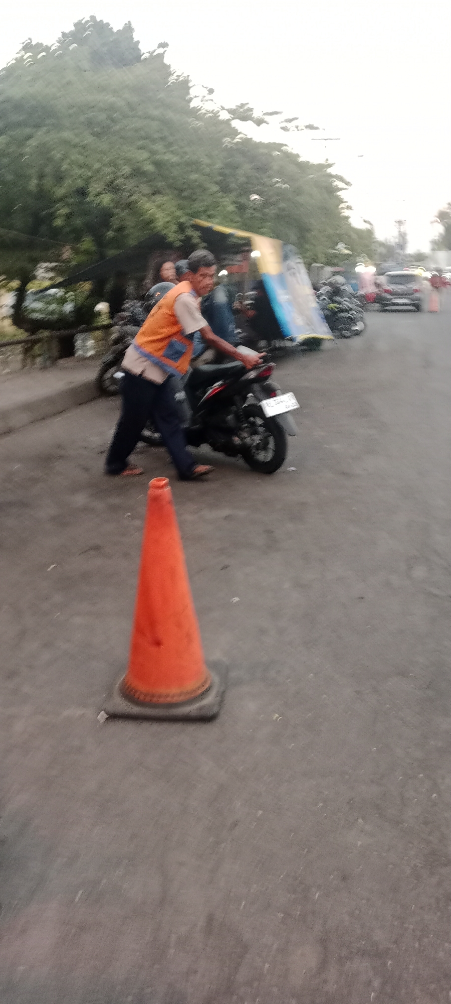 The image depicts a street scene in Ngemplak, Indonesia. In the foreground, a bright orange traffic cone stands on the asphalt. Behind the cone, a man wearing a light-colored shirt and a vibrant orange vest is pushing a dark-colored motorcycle. He is looking towards the viewer with a neutral expression. Several other motorcycles are parked or present in the background, along with some trees and structures that suggest an urban or semi-urban setting. The sky appears overcast or hazy. The overall lighting suggests it might be late afternoon or early morning.