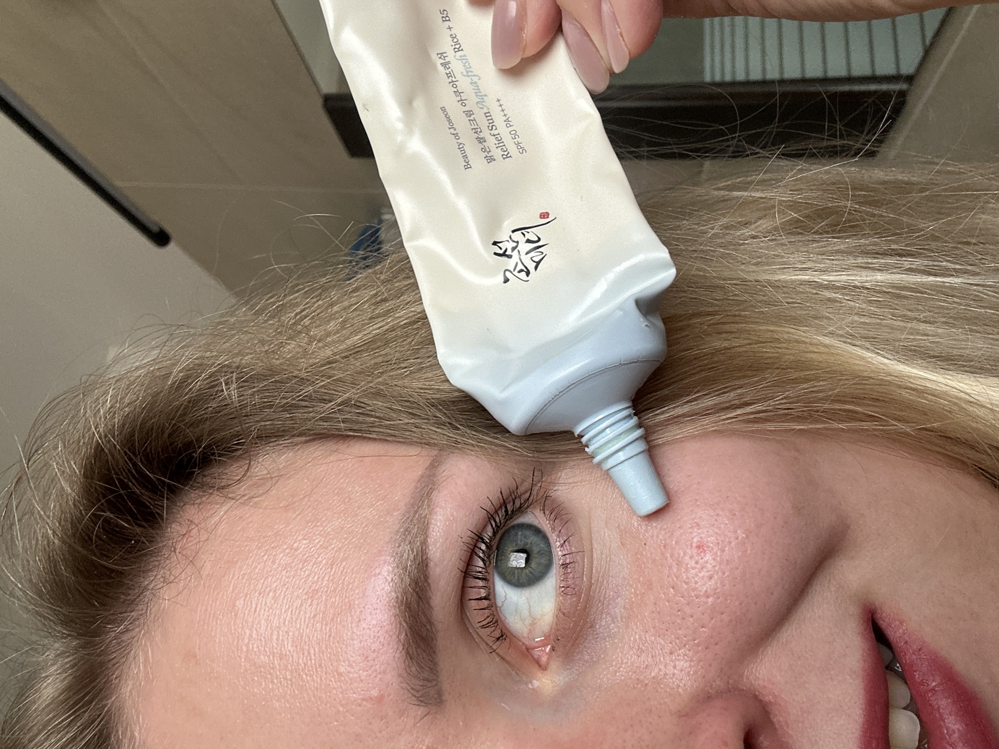 Here is a detailed description of the image:
The image shows a close-up, angled shot of a woman's face, positioned upside down in the frame. A tube of "Beauty of Joseon" Relief Sun Aqua-fresh Rice + B5 SPF50 PA++++ sunscreen is held above her face, with the tube's nozzle positioned directly over her eye. The woman has blonde hair and fair skin. Her eye is open, and you can see her eyelashes and eyebrows. A small red mark is visible on her cheek near the tube's nozzle. Her mouth is partially visible, showing her teeth and lips.
The background appears to be a light-colored wall, possibly a bathroom setting. The location, Petah Tikva, Israel, cannot be determined from the visual information provided in the image.
