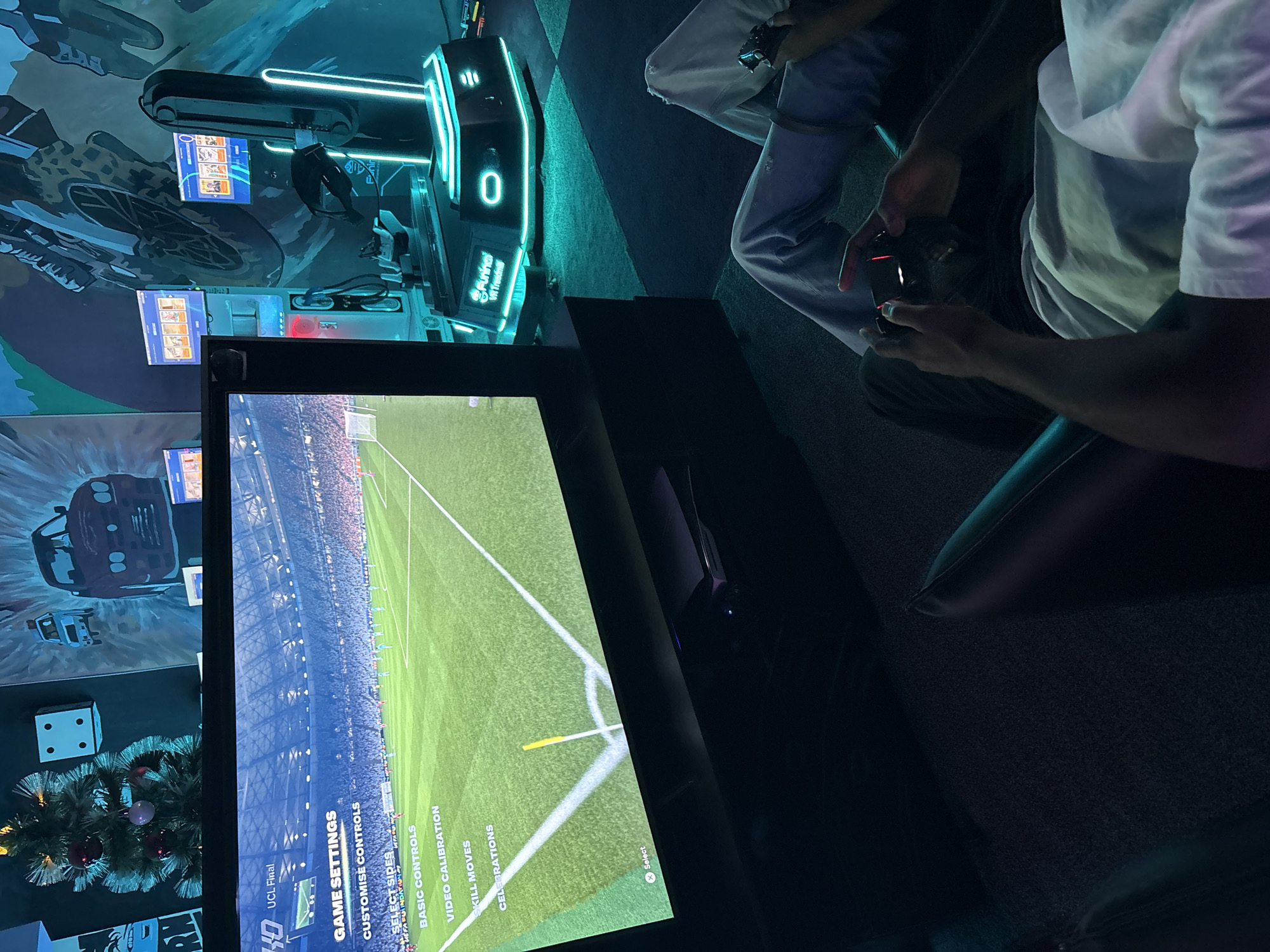 The image captures an indoor gaming or entertainment center in Abuja, Nigeria, featuring two partially visible individuals engaging with gaming equipment. One person, wearing a white t-shirt and light-colored pants, is seated and engrossed in a video game, holding a black and red controller. The game, displayed on a large flat-screen monitor, appears to be a soccer (football) match, showing a stadium interior and a green field. The monitor's menu displays "UCL Final" and "GAME SETTINGS" with options like "CUSTOMISE CONTROLS," "SELECT SIDES," "BASIC CONTROLS," "VIDEO CALIBRATION," "SKILL MOVES," and "CELEBRATIONS." An "X Select" prompt is also visible at the bottom right of the screen.
In the background, a "Funinai VR Treadmill" with prominent neon blue lighting and a digital display showing "0" is visible. Other gaming machines or arcade setups with smaller screens are also present. The walls are adorned with large, cartoonish murals of cars and mechanical elements, contributing to a futuristic or action-themed ambiance. A small, decorated Christmas tree with red and silver ornaments, and a large white dice decoration, suggest a festive or playful atmosphere. The overall lighting is dim, accentuated by the glowing neon lights of the machines, indicating an evening or night setting. The floor features a dark, geometric pattern.