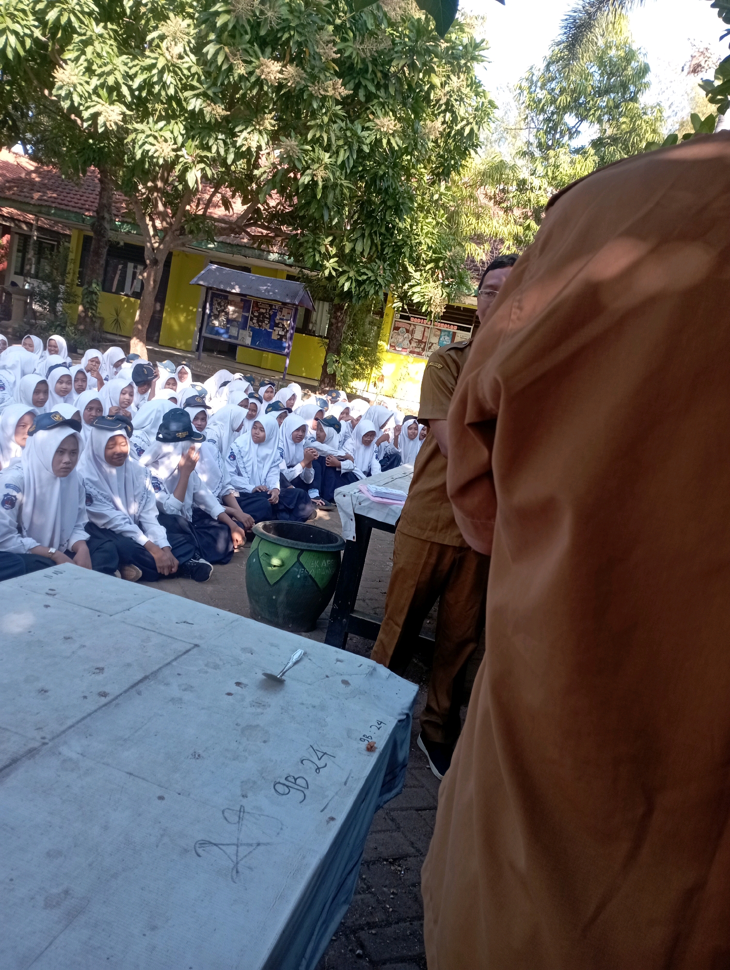 In Banyuurip, Indonesia, a group of uniformed students are gathered outdoors, sitting on the ground in front of a table. The students are predominantly female, wearing white hijabs and uniforms, and appear to be listening to an adult figure in a brown uniform, partially visible in the foreground. The setting appears to be a schoolyard, with trees, a building with yellow walls, and a bulletin board visible in the background. A large green pot is positioned near the students, and the table in front of them has some writing on it, including "9B 24". The lighting suggests it is daytime.