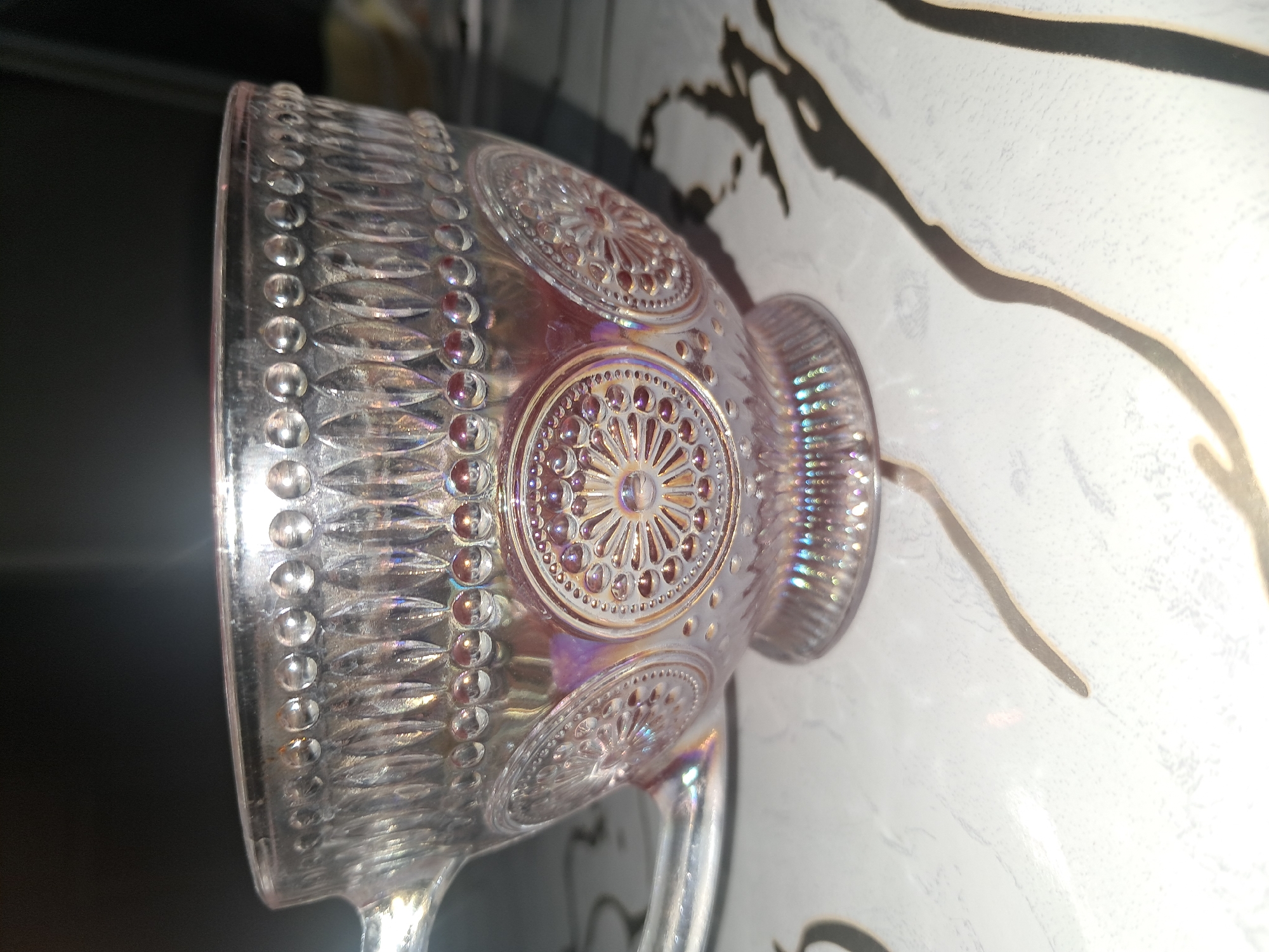 This media file shows a close-up, angled shot of a clear glass cup with iridescent qualities. The cup is ornately decorated with raised circular patterns resembling sunbursts, each surrounded by a border of small, raised dots. Vertical ridges are also visible along the exterior of the cup, and the glass has a pearlescent sheen that reflects a spectrum of colors, including pink, purple, and yellow. The cup appears to be empty and is positioned on a surface that has a white, marbled pattern with dark grey, abstract shapes. The background is dark, suggesting an indoor setting with artificial lighting that contributes to the iridescent effects on the glass. The focus is sharp on the cup's details, while the background is blurred. No people or text are visible in the image.