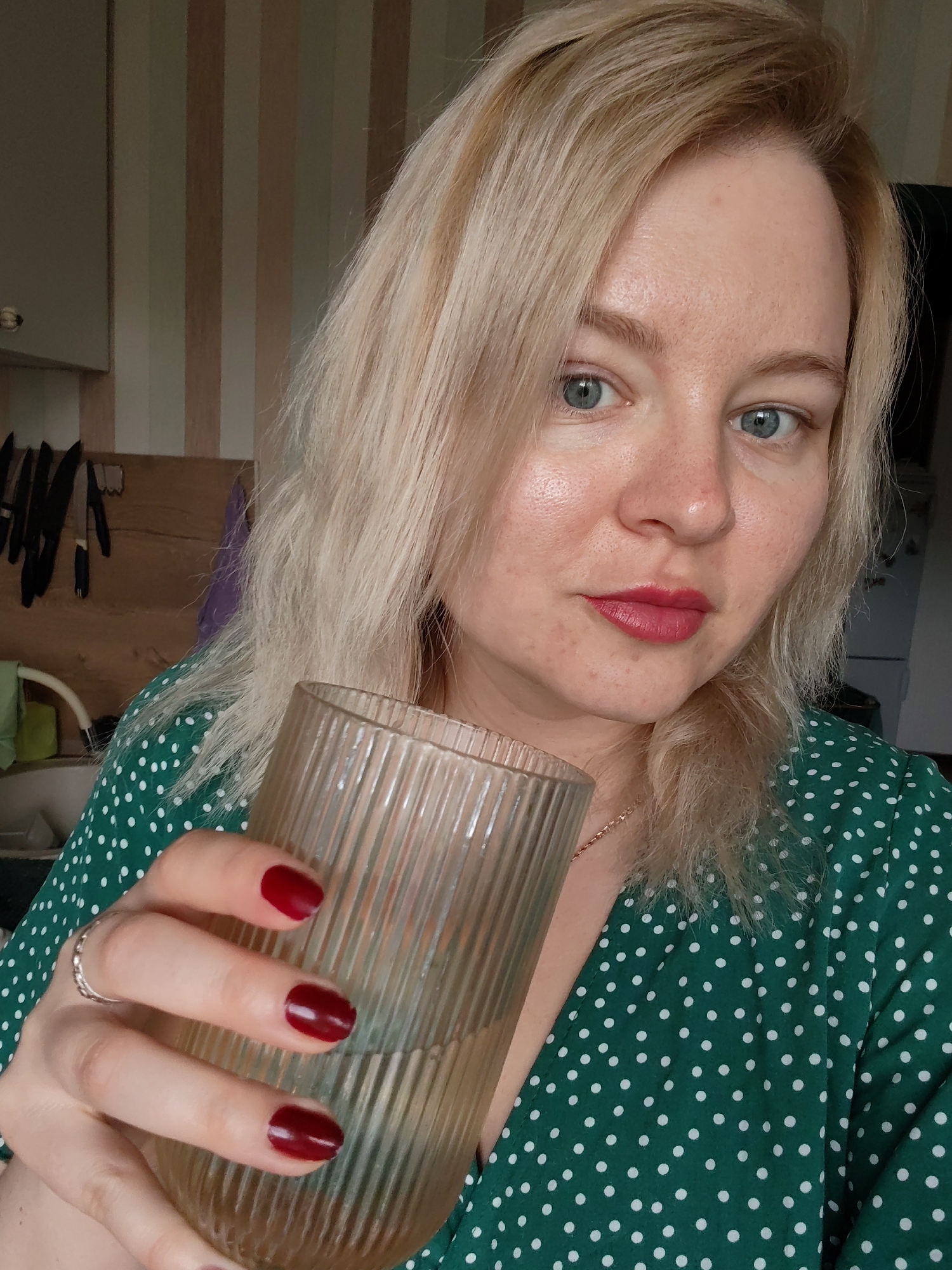 A woman with blonde hair and blue eyes is holding a glass of water in her right hand. She has red lipstick and matching nail polish. She is wearing a green dress with white polka dots. The background includes a kitchen area with cabinets, knives hanging on the wall, and striped wallpaper. Based on the available information, there's nothing to suggest that the photo was taken in Izhevsk, Russia.