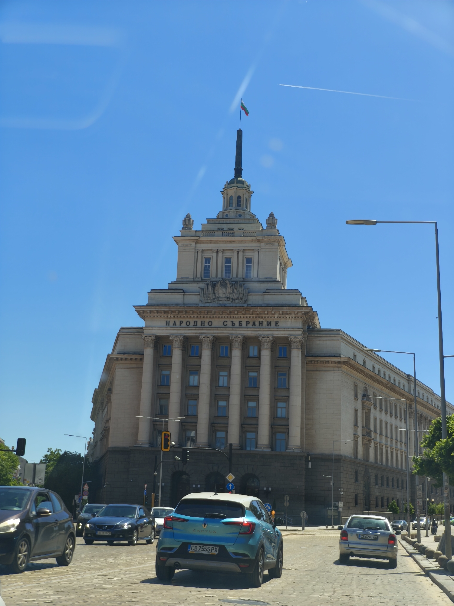 The image shows the National Assembly building in Sofia, Bulgaria, during daylight. The building is in the foreground, with cars driving on a cobblestone street in front of it. The sky is blue with a contrail from an airplane. A Bulgarian flag flies atop the building. There are lampposts and trees along the street. Traffic lights are visible.