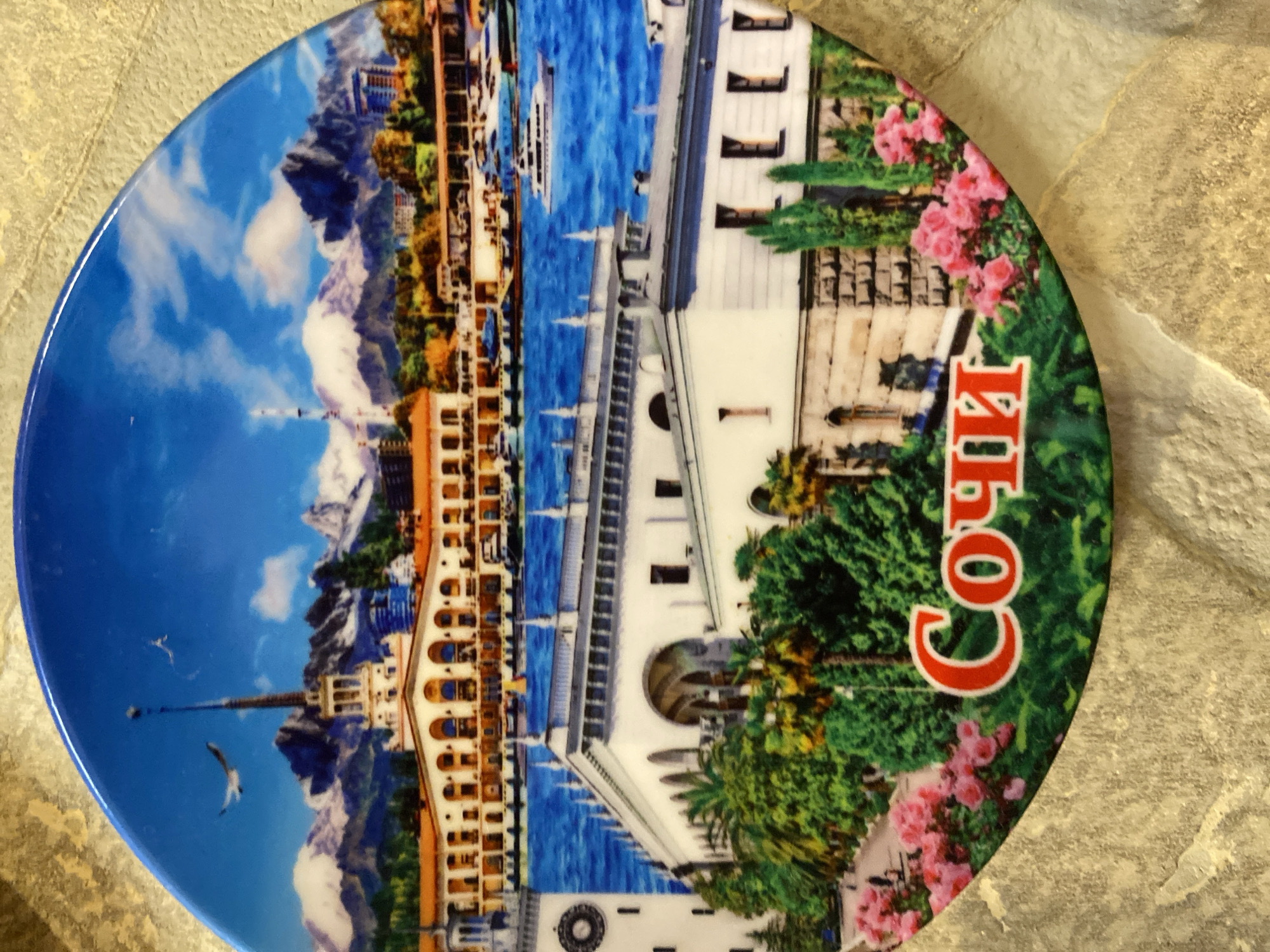 This image shows a circular souvenir plate with a scenic depiction of Sochi, Russia. The plate features a vibrant landscape with a blue sky filled with white clouds and distant snow-capped mountains. In the foreground, there is a large white building with arched windows, surrounded by lush green trees and blooming pink flowers. A body of water, likely the sea, is visible behind the building, with several yachts and boats docked. The plate also displays text in Russian, which reads "Сочи" (Sochi). The plate is resting on a textured, light-colored surface that appears to be stone or tile. The image is a close-up, focusing on the details of the plate's design.