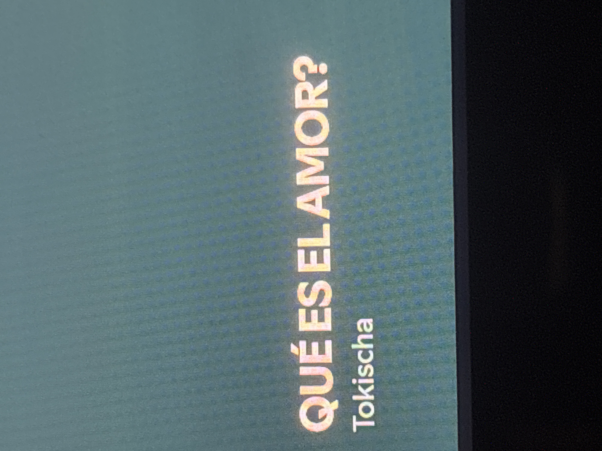 The image shows a vertical text display on a screen. The text reads "QUÉ ES EL AMOR?" above "Tokischa." The background is a textured teal color, and a black border is visible on the right side of the image.