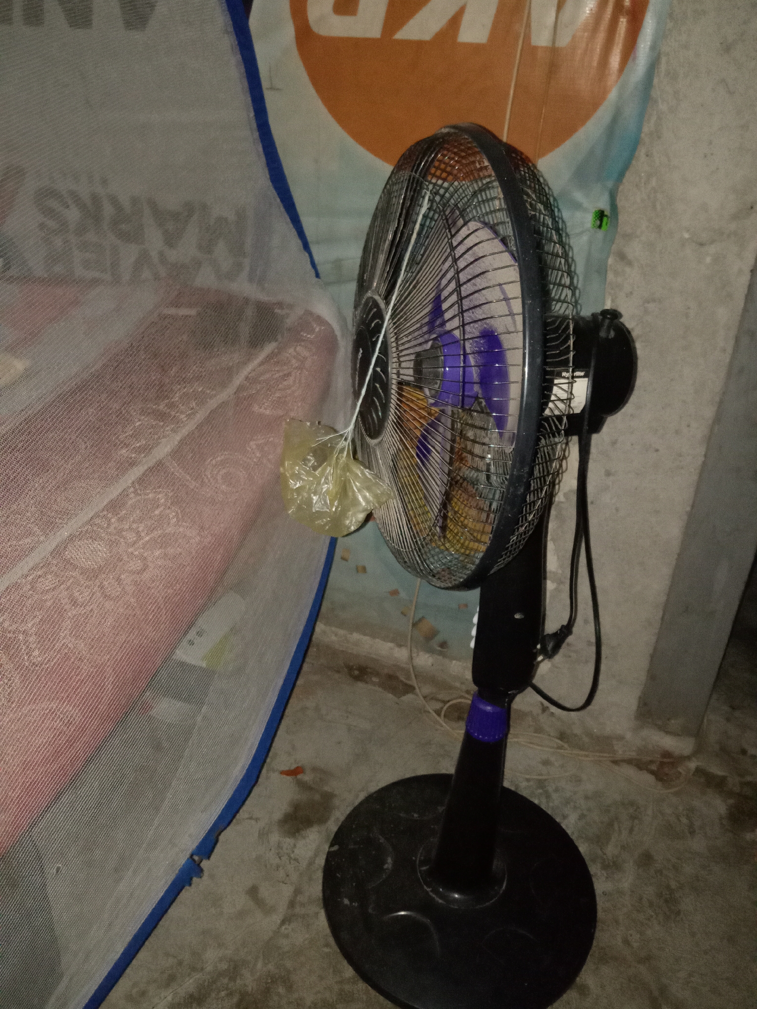 The image depicts an indoor scene, likely a bedroom, in Jumput, Indonesia. A black oscillating fan stands on a concrete floor, its blades partially visible behind a metal grill. The fan is positioned next to a mattress covered with a pink blanket and a white mosquito net. Behind the fan, a light blue and orange banner hangs on a concrete wall. The overall lighting suggests it is nighttime or a dimly lit room.