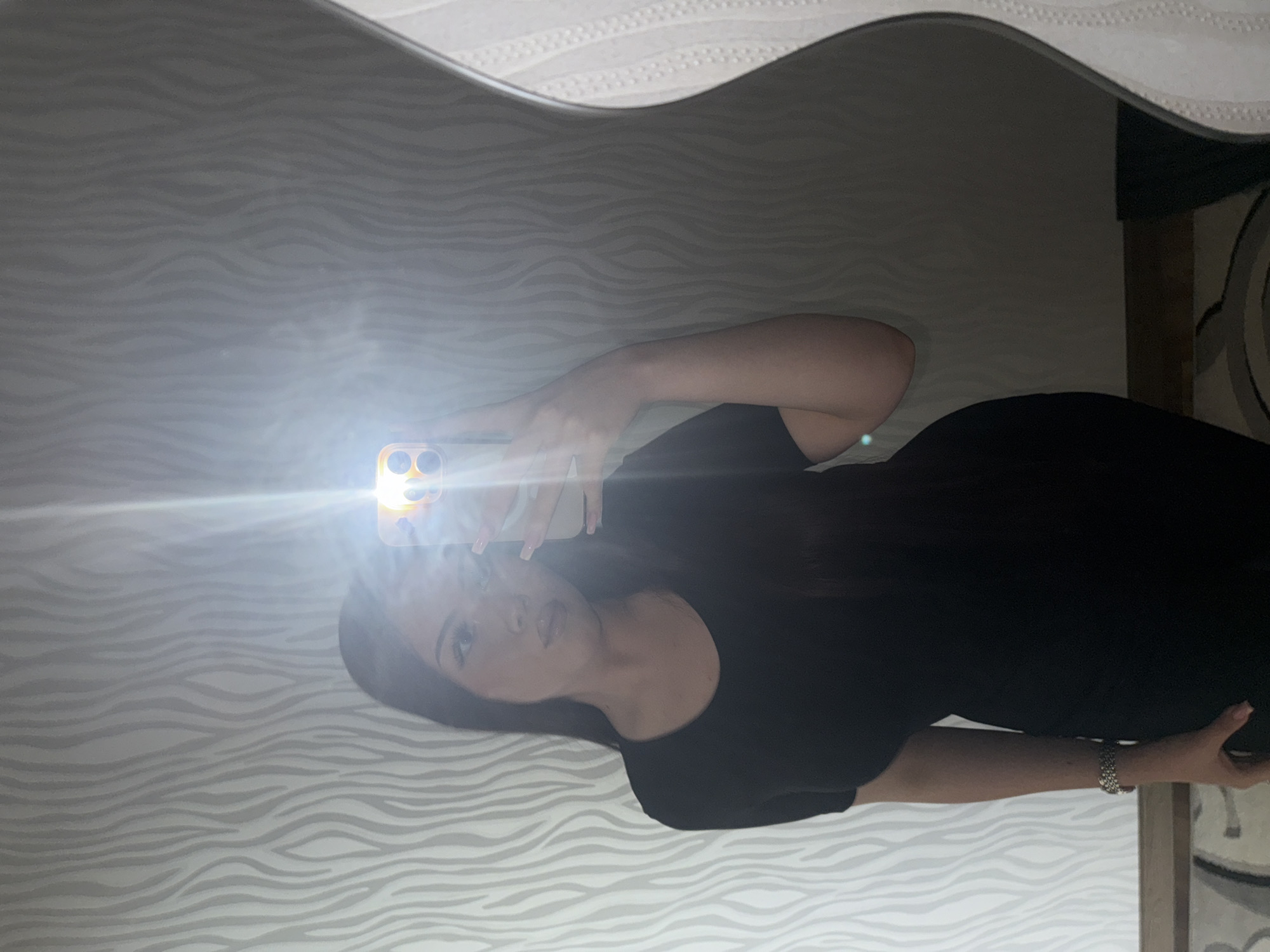 A woman is taking a selfie in a mirror. The mirror reflects a room with patterned wallpaper and part of a rug. The woman is wearing a black top and a bracelet. Her location cannot be determined to be Naberezhnye Chelny, Russia, as there is no discernible information in the image to suggest that location.