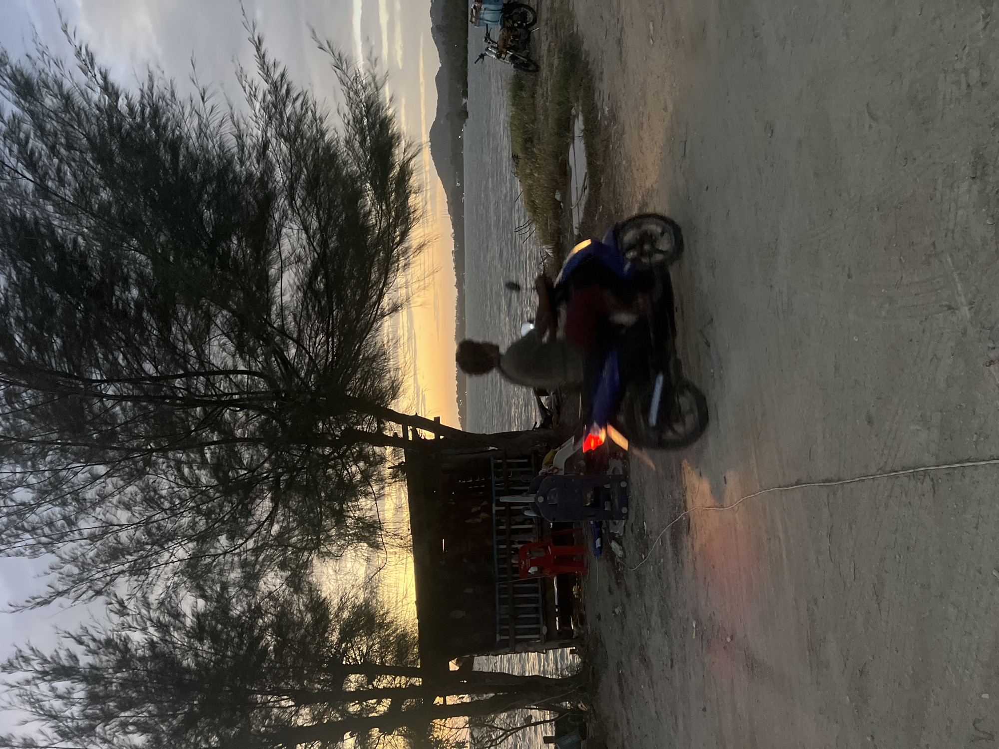 At dusk on the coast of Ko Pha-ngan District, Thailand, a person rides a blue motorcycle from left to right in the foreground. The rider, facing away from the camera, is partially obscured by the motion blur of the motorcycle. The motorcycle's taillight is illuminated, casting a red glow on the dusty ground. In the middle ground, a small, weathered wooden shack with a corrugated metal roof stands near the water's edge. Two figures are seated on chairs outside the shack. The shack is flanked by sparse trees with thin, dark foliage. The sea stretches out to the horizon, where a hilly landscape is visible under a pale, cloud-streaked sky. Another motorcycle, a lighter color, is parked on the grassy embankment to the right of the shack, with a blue cooler bag strapped to its rear. The ground is predominantly dirt and sand, with some scattered debris.