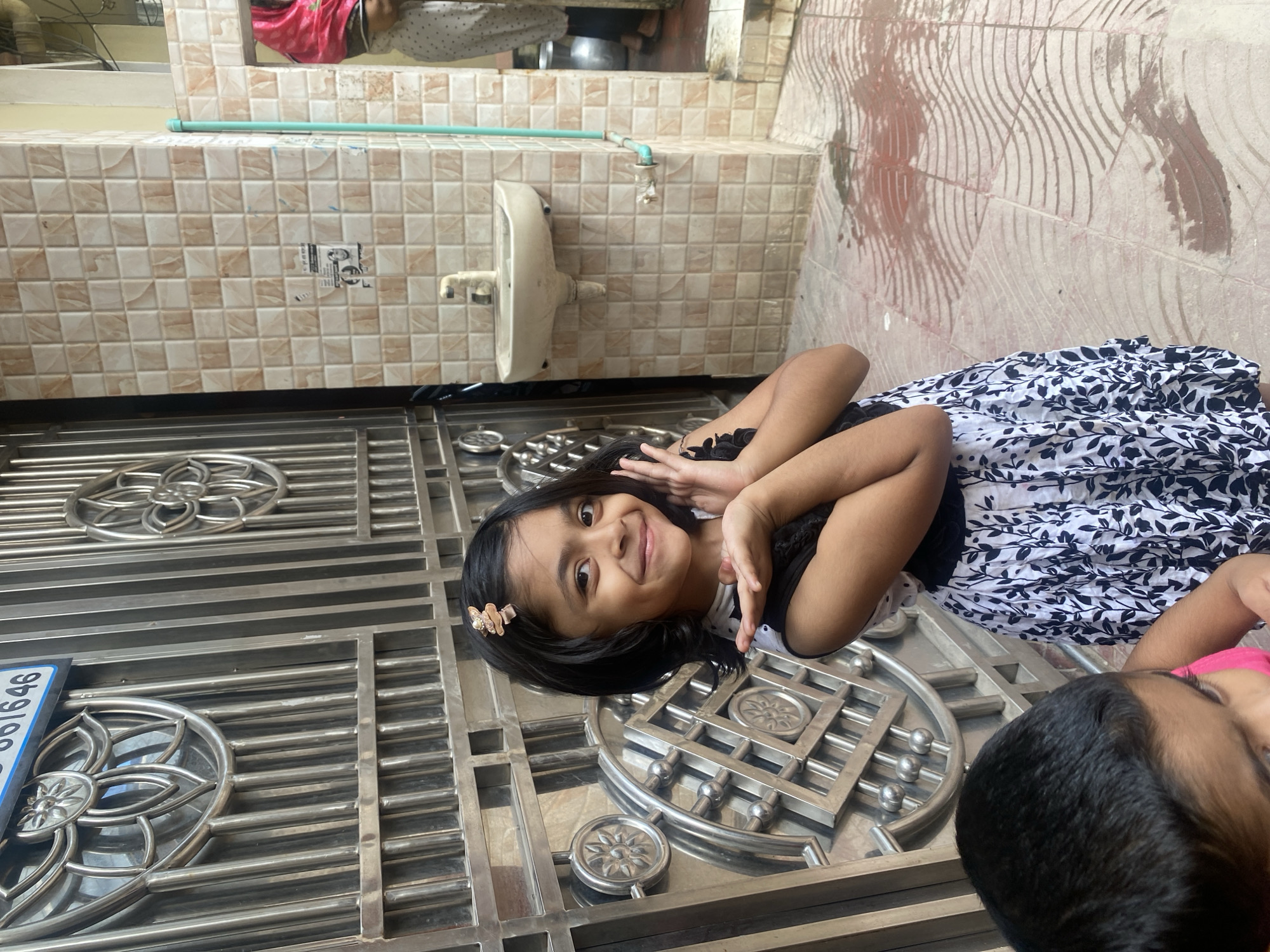 A young girl with dark hair and a butterfly clip smiles at the camera, holding her hands up near her face. She is wearing a black and white floral dress. She stands in front of a silver metal gate with decorative circular designs. To the right of the gate is a tiled wall with a white sink and faucet. Another child, seen from behind, is in the foreground on the left. The ground is paved with tiles and appears wet in some areas. The scene is likely outdoors, possibly in a residential or public area.