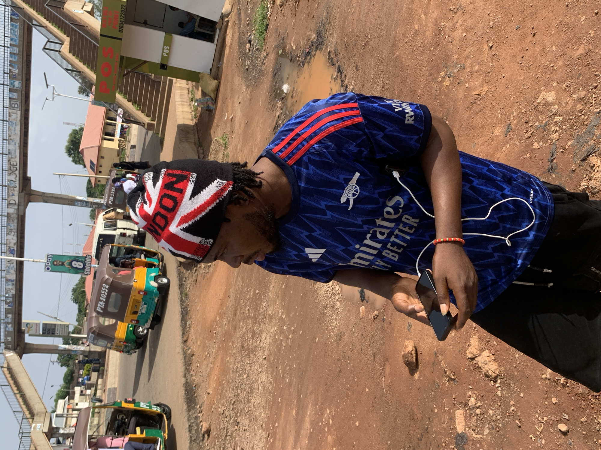 In Gura Top, Nigeria, a man stands in a dusty, outdoor environment, looking to his right. He wears a dark beanie adorned with the Union Jack flag and a blue short-sleeved shirt with red stripes on the shoulders and an Arsenal FC logo. The shirt also features white text, partially obscured, that includes "emirates" and "BETTER." He is holding a black smartphone, connected to a white cable that runs into his shirt. A red and white beaded bracelet is visible on his right wrist. The ground is a reddish-brown dirt with scattered stones and a small puddle. In the background, a pedestrian bridge arches overhead, with various signs and structures visible beneath it. Several auto-rickshaws, one green and yellow with "PTA-10552" visible, are parked or moving on a paved surface. A white van and a green and yellow building with solar panels are also in the distance. A "POS" sign is visible on a green booth with a staircase leading up to it.