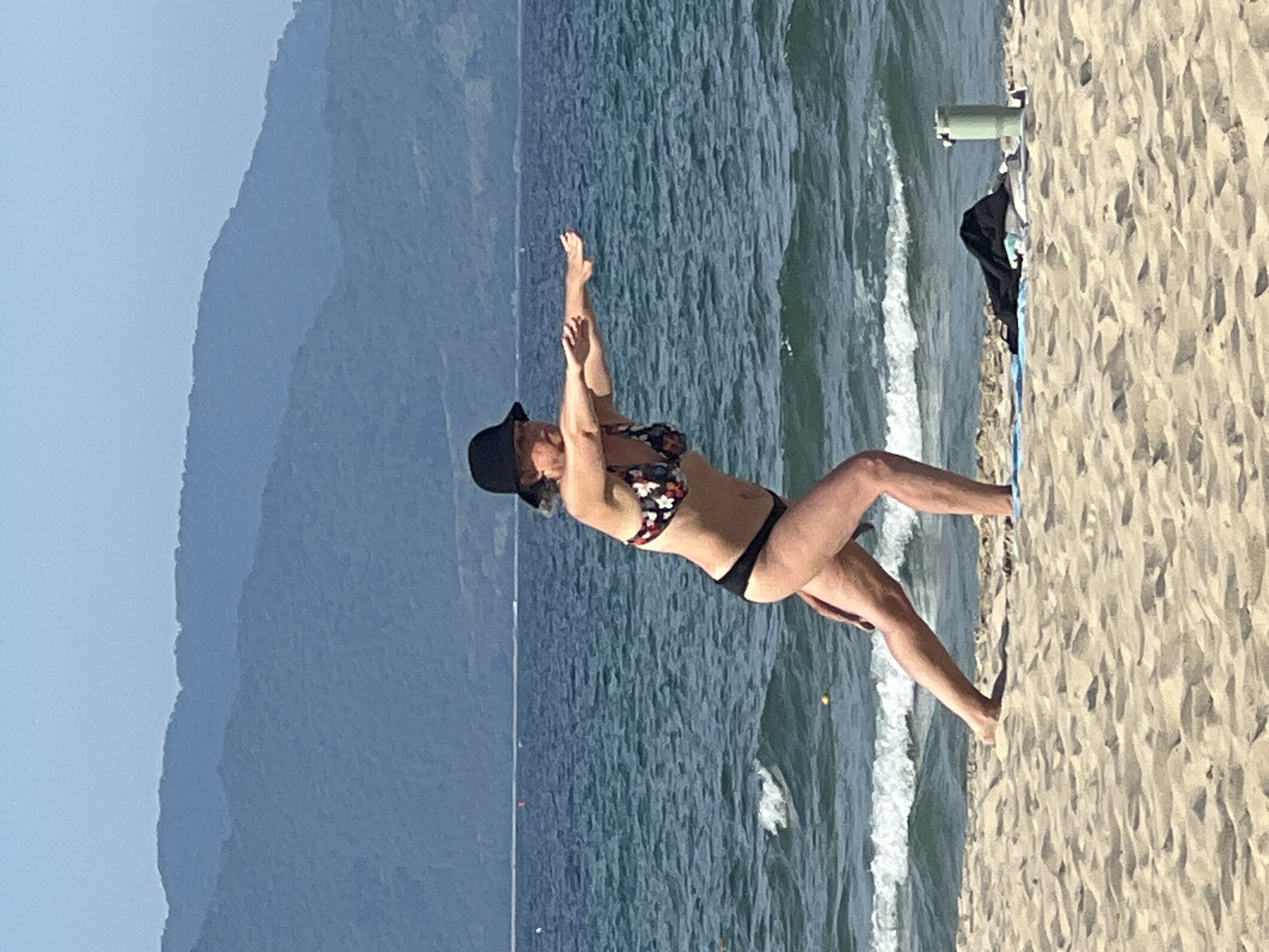 A woman in a floral bikini top and black bottoms, wearing a black bucket hat, performs a yoga pose on a sandy beach. She is facing to the right of the frame, with her left leg forward and bent, and her right leg extended behind her. Her arms are outstretched forward, parallel to the ground. The ocean, with moderate waves, is behind her, and in the distance, there are layered mountains under a clear blue sky. On the sand near her, there is a blue towel, some clothing, and a green water bottle. The scene is consistent with a location along the coastline of Đà Nẵng, Vietnam, known for its beaches and mountainous backdrop.