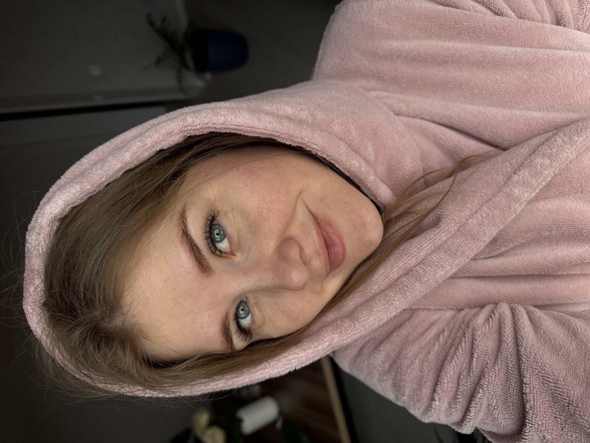 A woman is lying down, wearing a pink hooded robe. Her face is turned toward the viewer. She has light skin, light brown hair partially visible under the hood, and light blue eyes. The background is dark and indistinct, with a hint of a chair or other furniture visible. The photo appears to have been taken indoors. The location in Gornyi (Dvurechenskoe S/P), Russia is unsupported by the visual data.